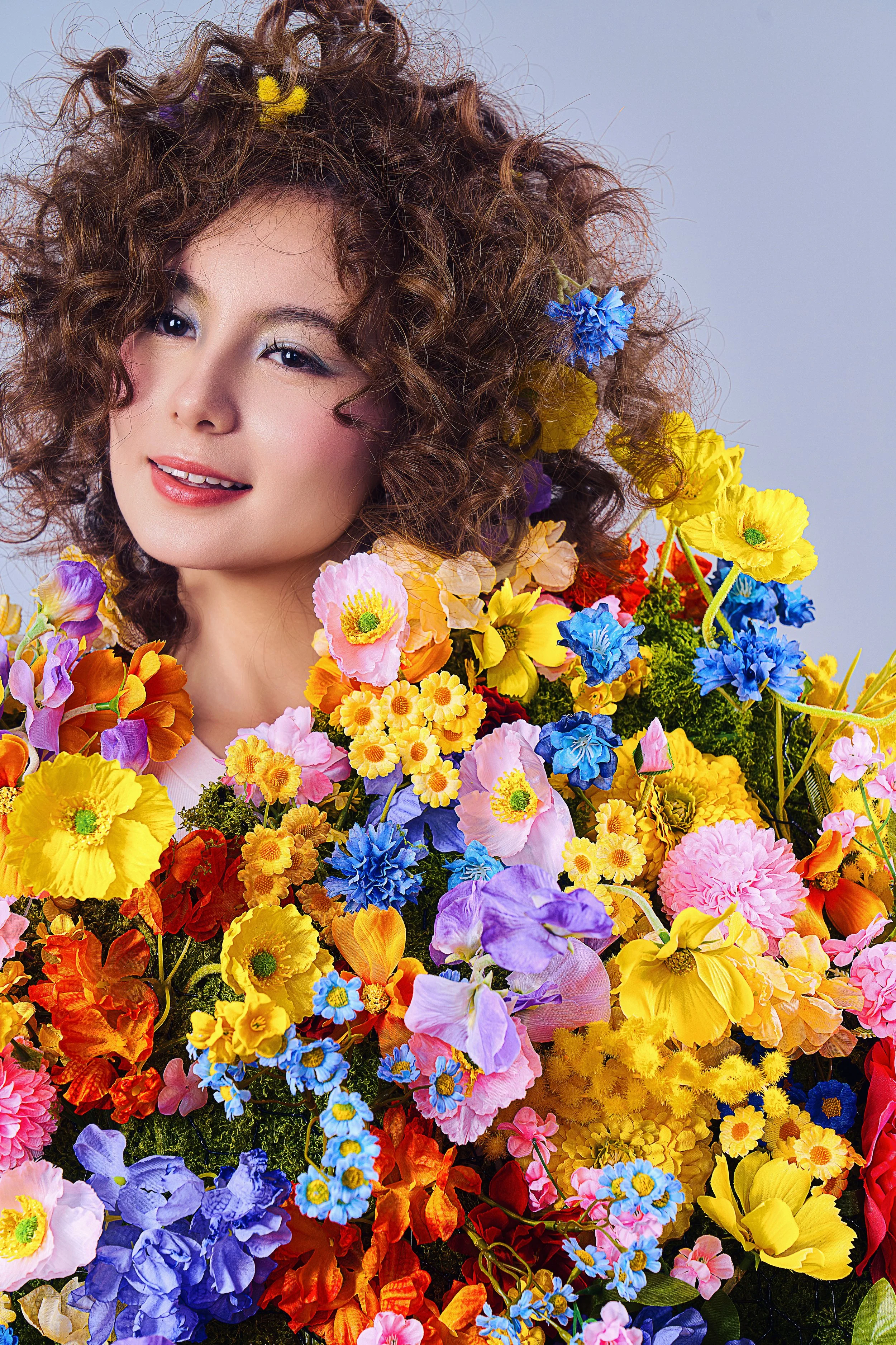 A woman with curly hair surrounded by a large variety of colorful flowers, including yellow, pink, purple, blue, and red blooms.