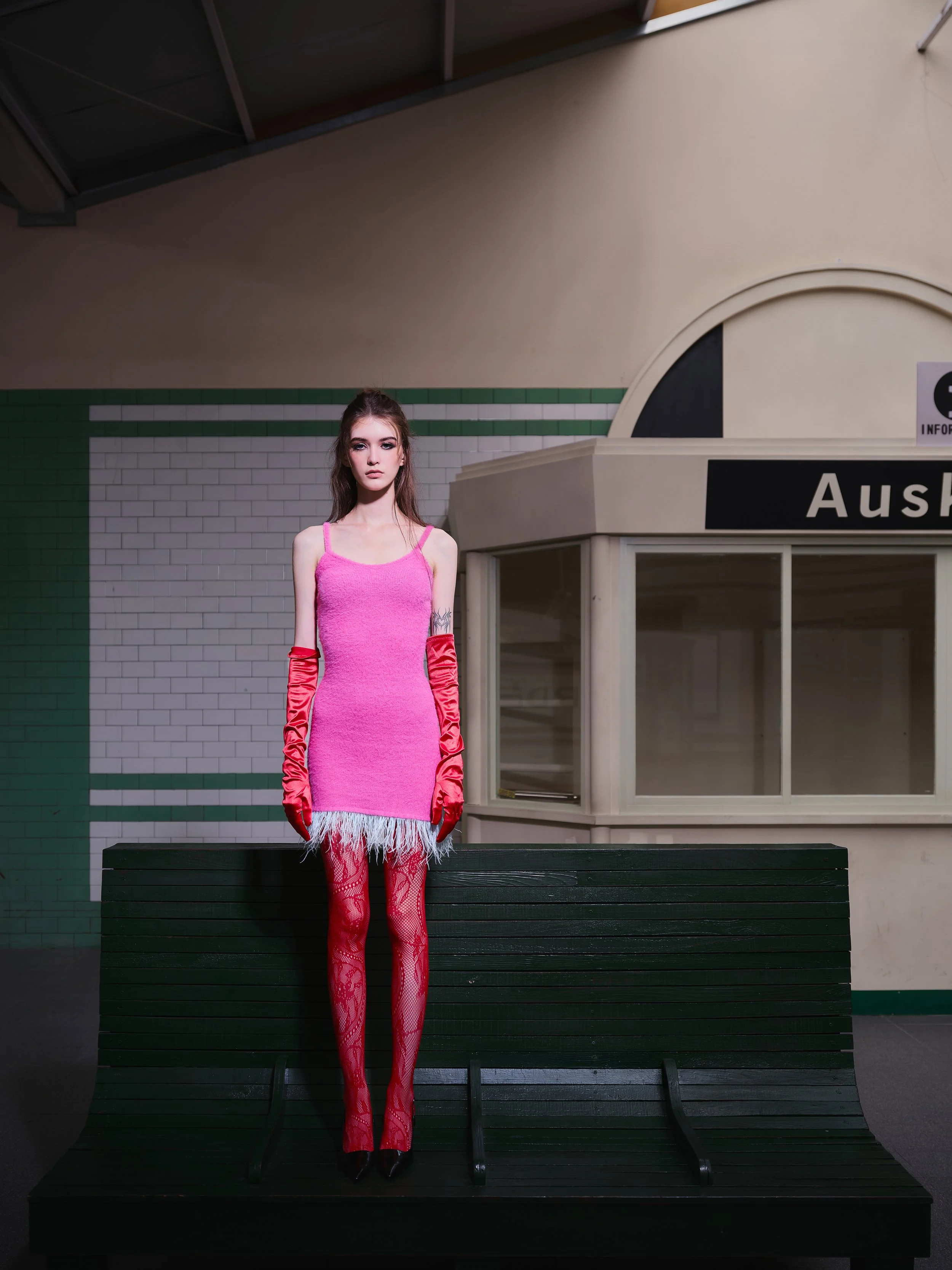 A young woman in a pink dress with feather trim, wearing red lace gloves and red lace tights, standing on a dark green bench in an indoor space that appears to be a train station or similar facility.