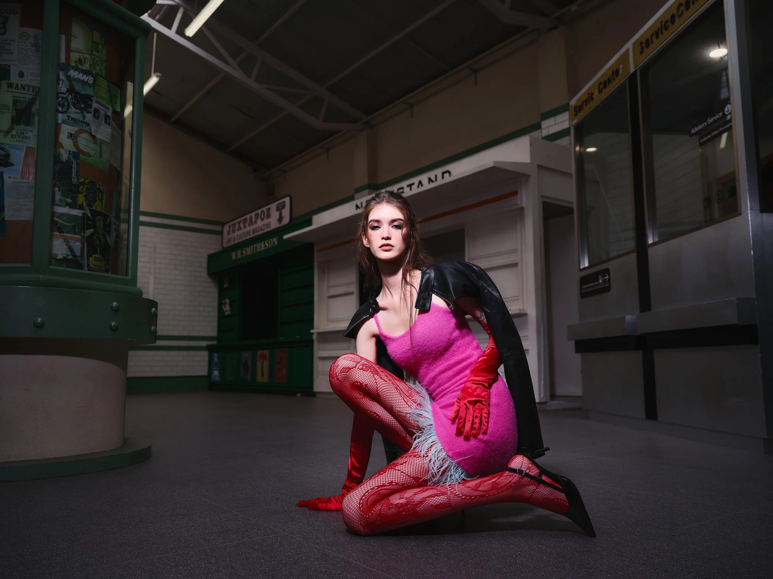 A young woman with long brown hair kneeling on the floor of a train station, wearing a pink dress, red fishnet stockings, red gloves, black high heels, and a black leather jacket draped over her shoulders. She has a serious expression and is looking 