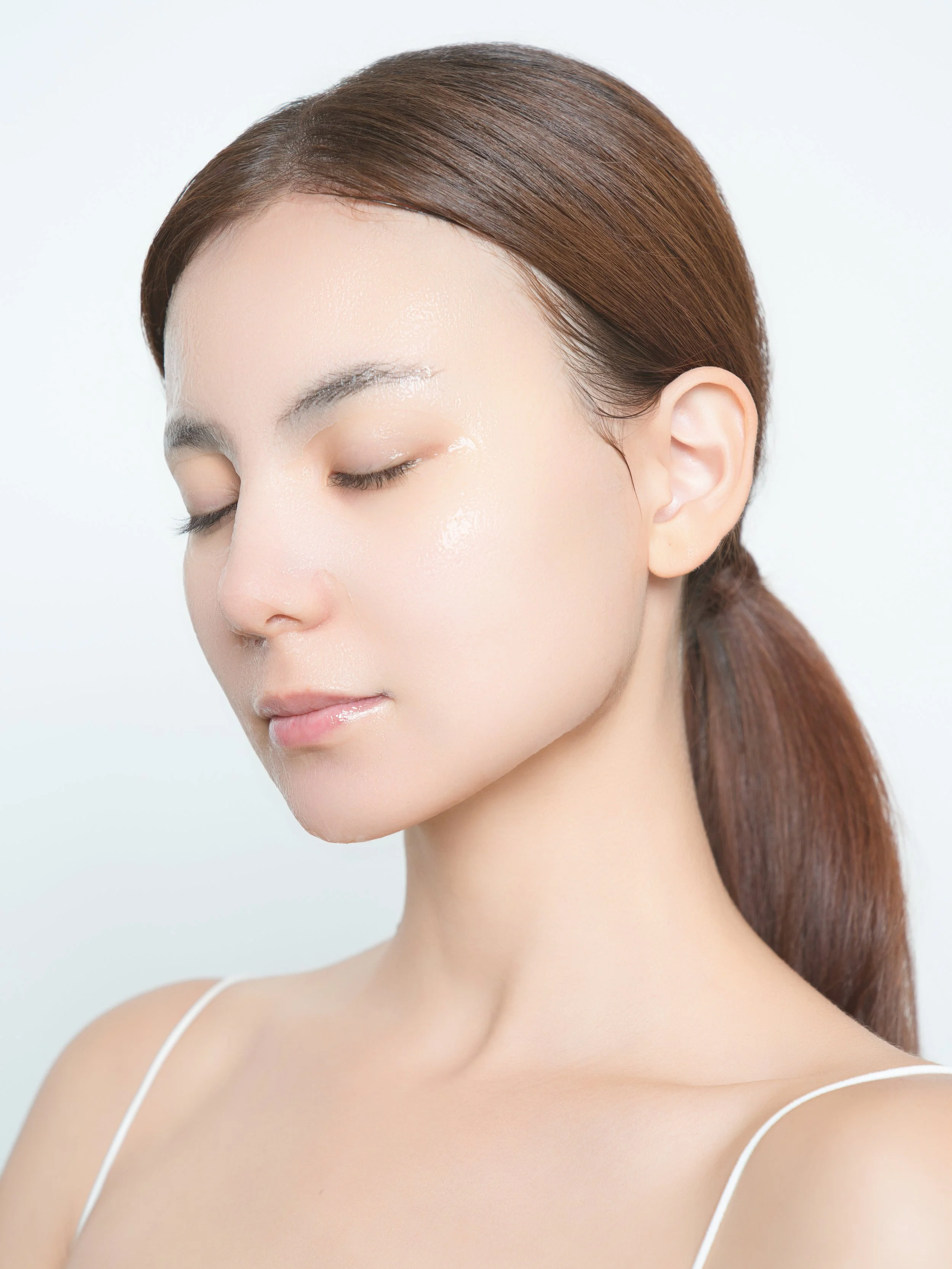 Close-up of a young woman with clear, smooth skin and closed eyes, with brown hair tied back, against a plain light background.