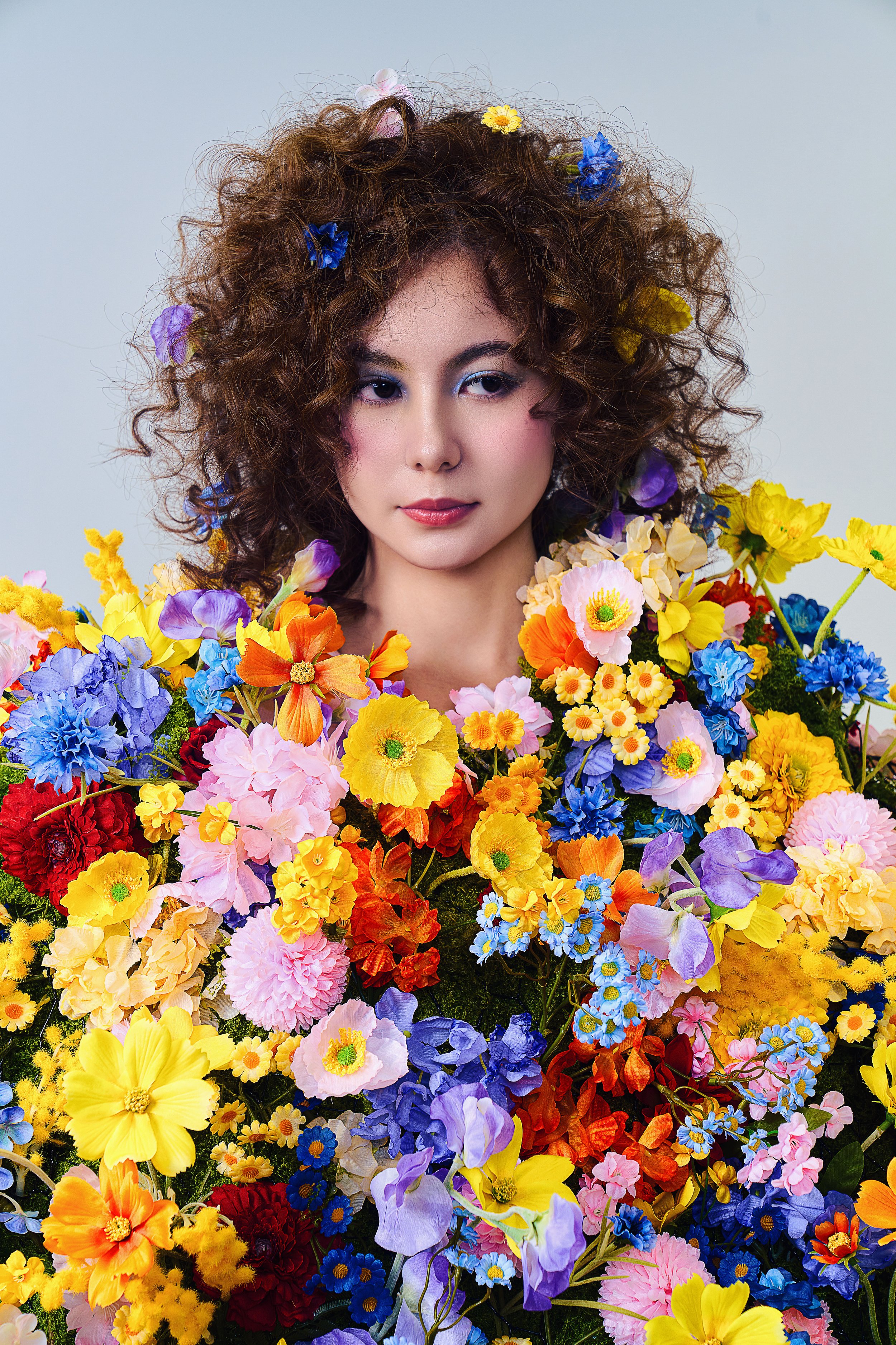 A woman with curly hair decorated with colorful flowers, surrounded by a large bouquet of bright and pastel flowers.