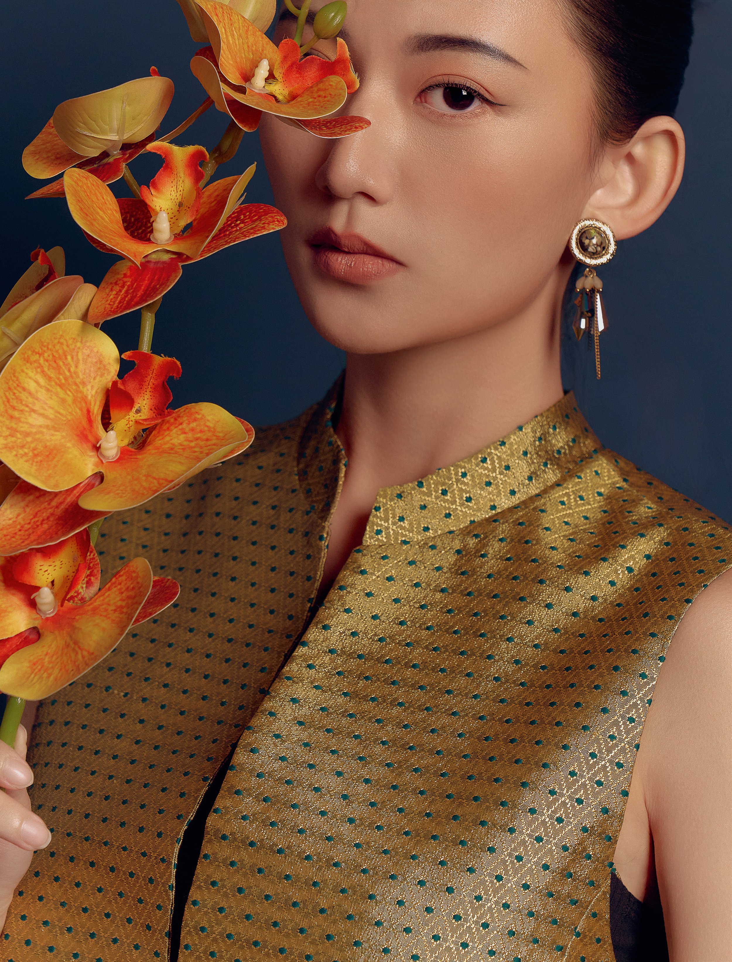 A woman with fair skin, dark hair, and subtle makeup, wearing a gold and teal polka-dotted sleeveless top and ornate earrings, holds a cluster of orange orchids close to her face against a dark background.