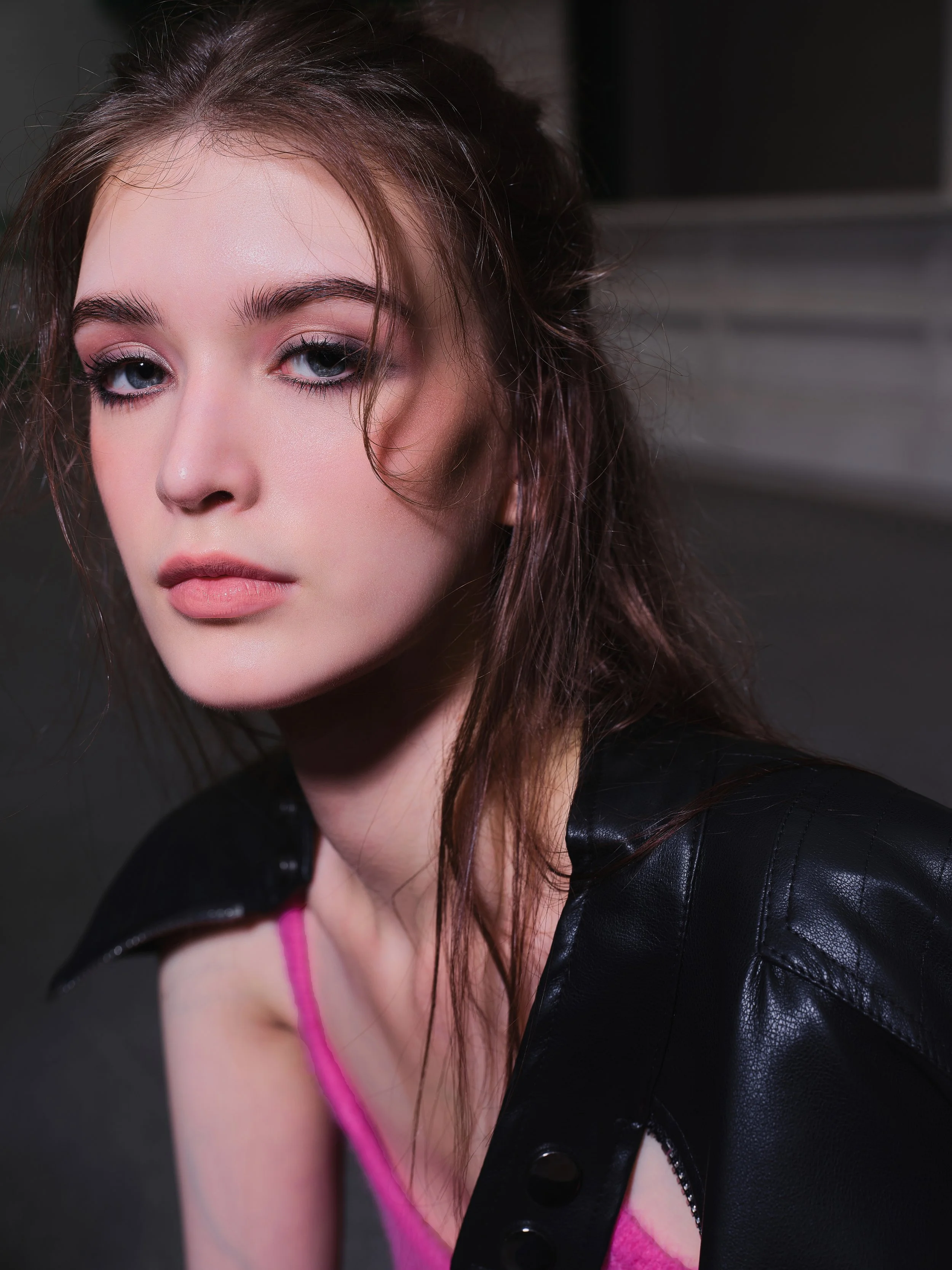 Close-up portrait of a young woman with fair skin, light makeup, slightly wet, messy brown hair, wearing a black leather jacket over a pink top, against a dark background.