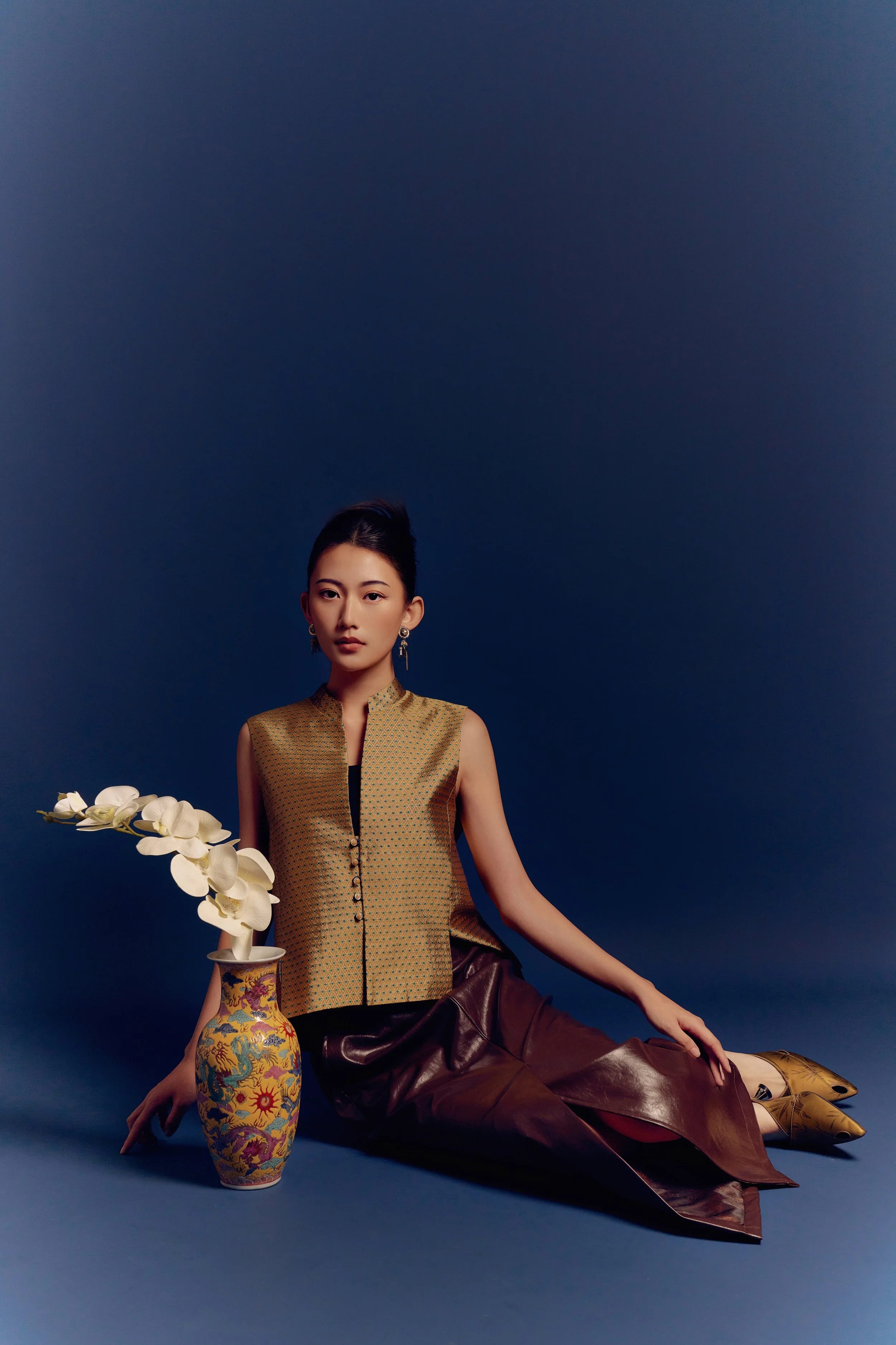 A woman sitting on the floor next to a vase with white orchids, posing against a dark blue background.