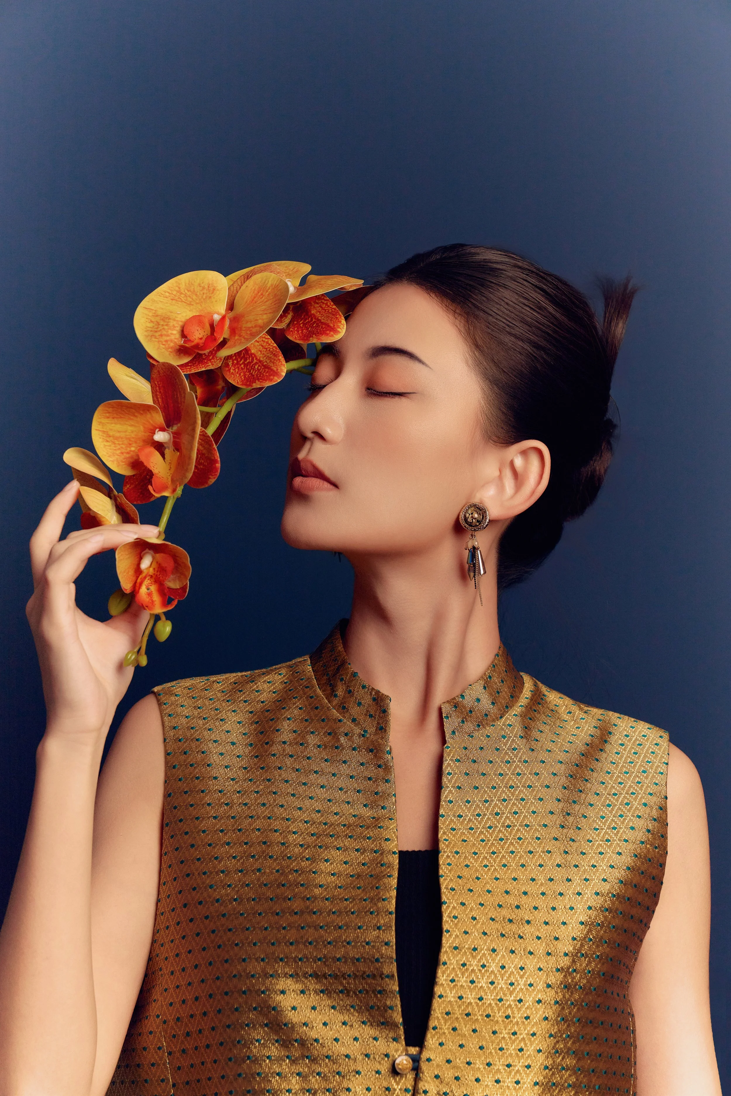 A woman with dark hair styled in an elegant updo, wearing a gold sleeveless top with small blue polka dots, earrings, and holding a branch of orange orchids near her face with eyes closed against a dark blue background.