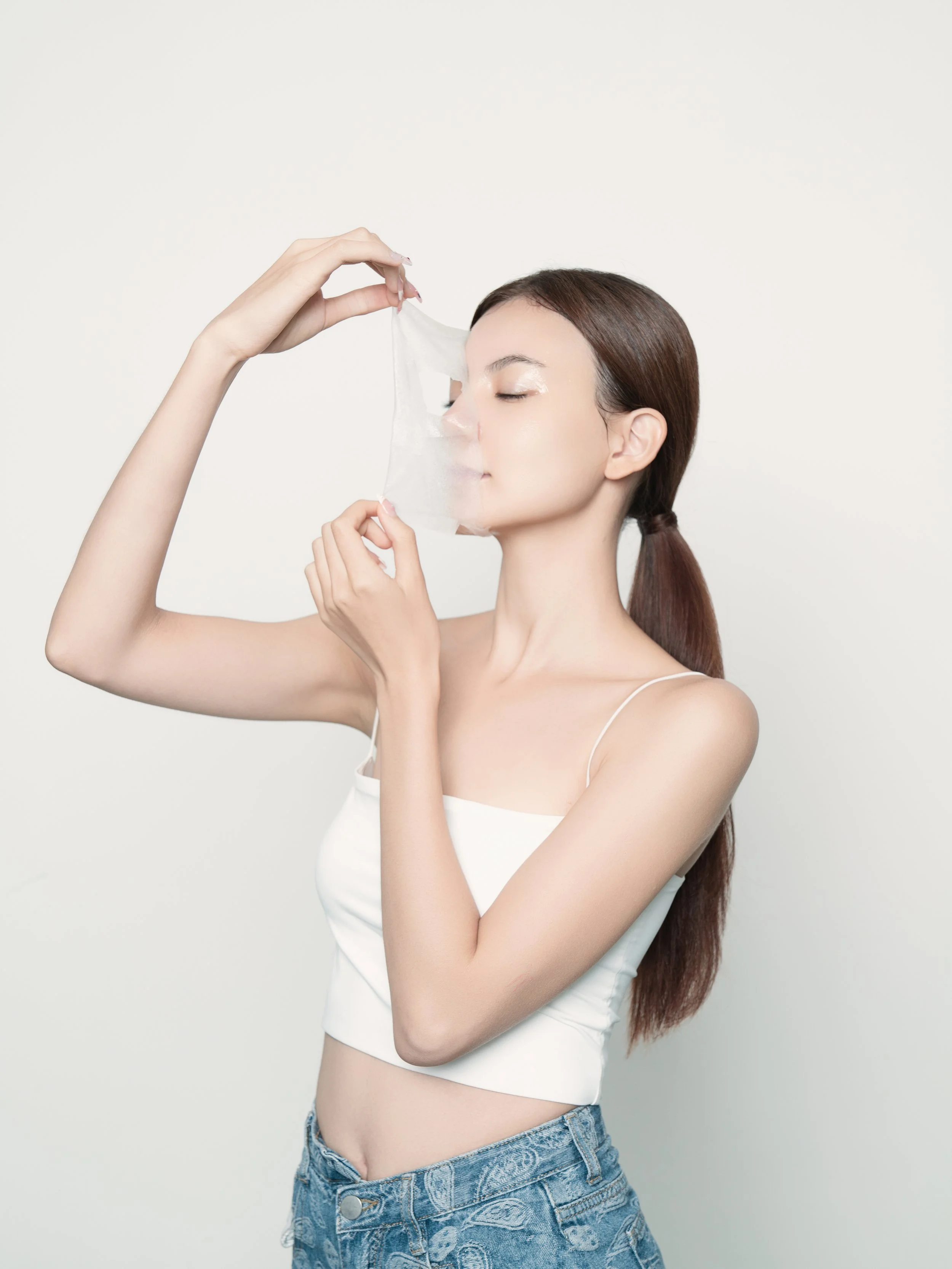 Young woman with long brown hair in a ponytail, wearing a white tank top and blue jeans, holding a cleansing wipe near her face with her eyes closed, standing against a plain white background.