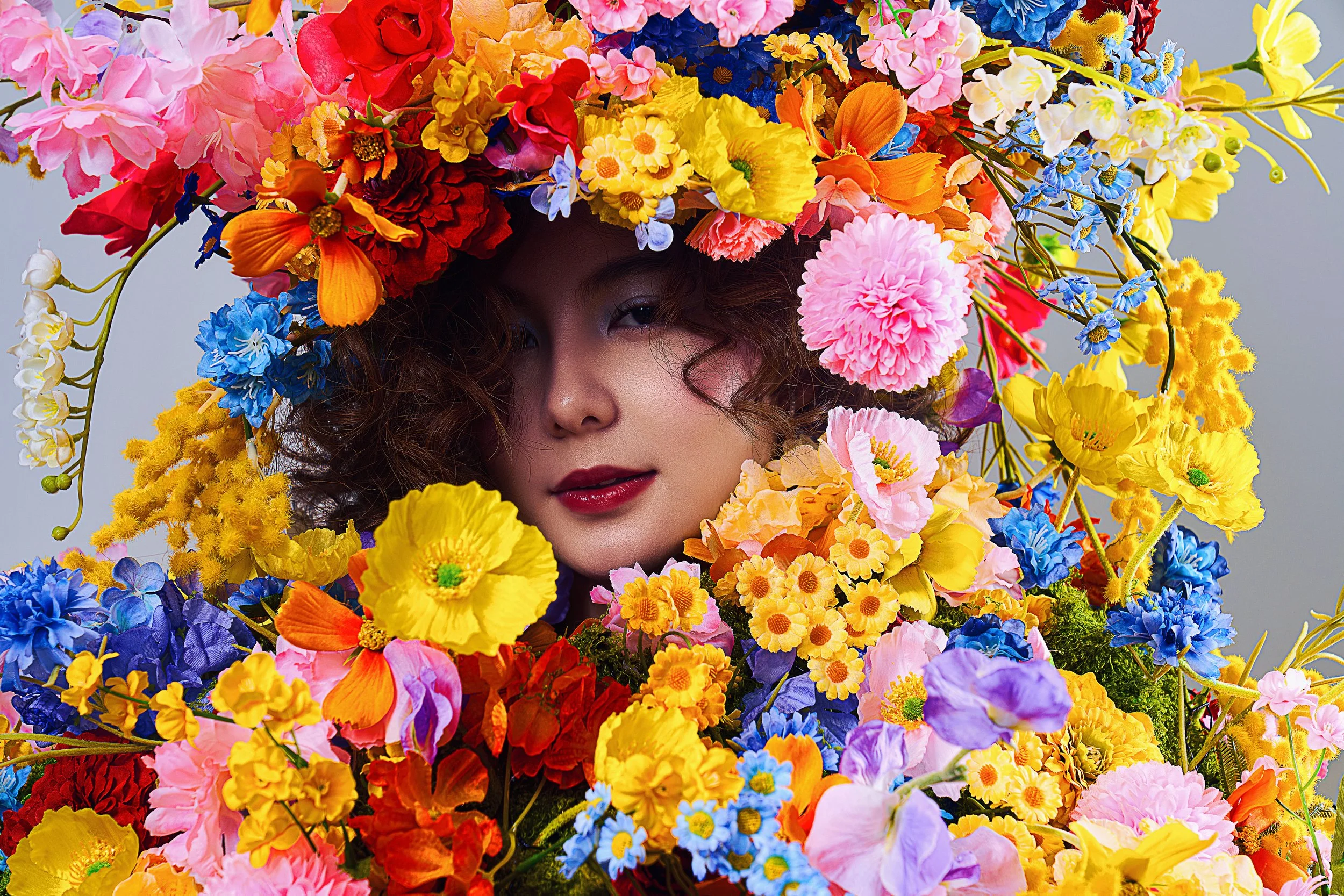 A woman with curly hair and red lipstick faces forward, surrounded by a colorful array of flowers including pink, yellow, orange, purple, and blue blooms, with a large floral headdress partly obscuring her face.