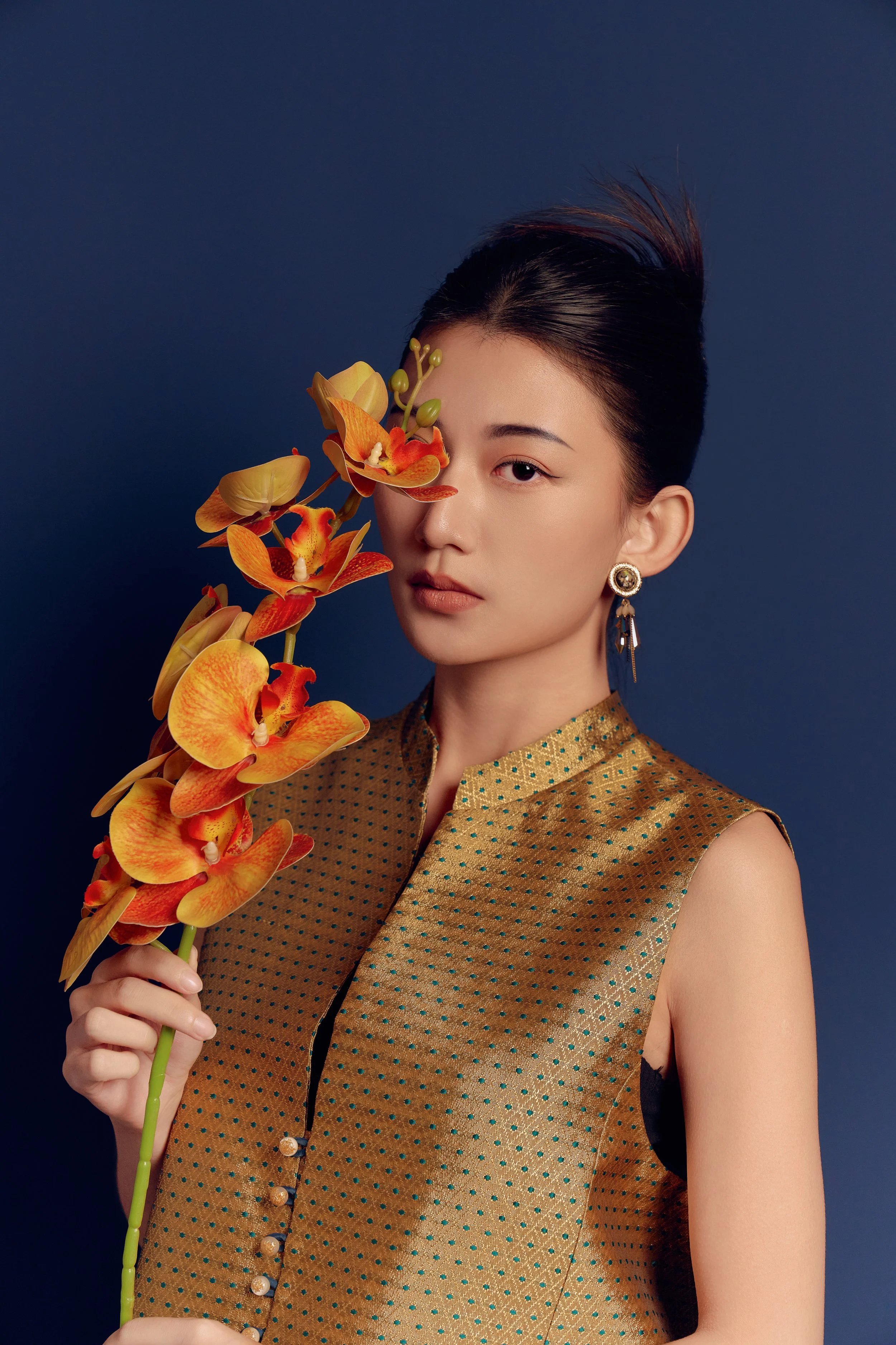 A young woman with dark hair styled in an updo, wearing a gold sleeveless top with small green polka dots, holding a stem of orange orchids, partially covering her right eye, against a dark blue background.