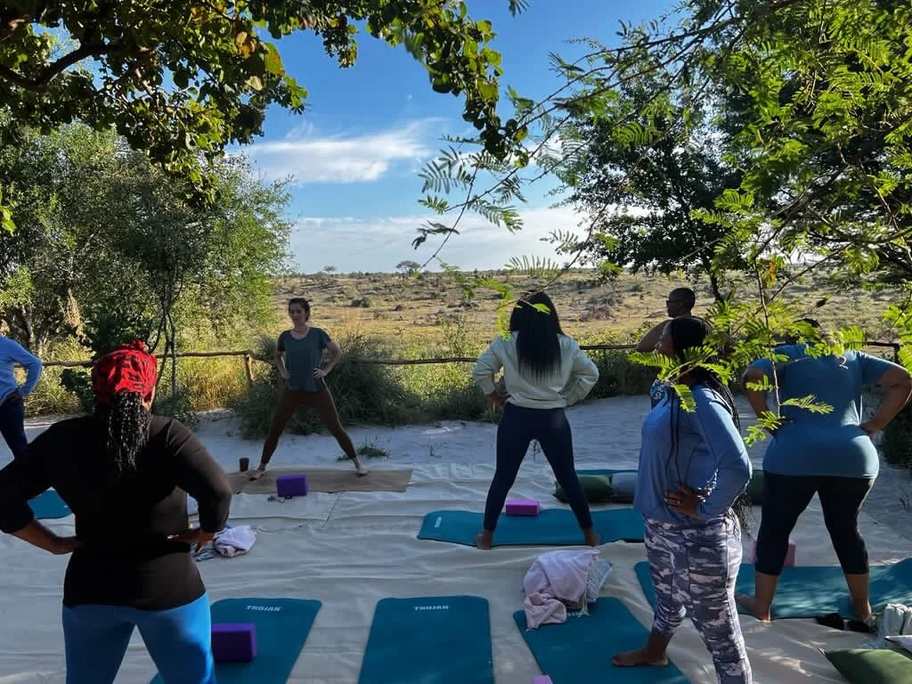 Women practicing yoga outdoors on mats in a shaded area with trees, with a landscape and clear sky in the background.