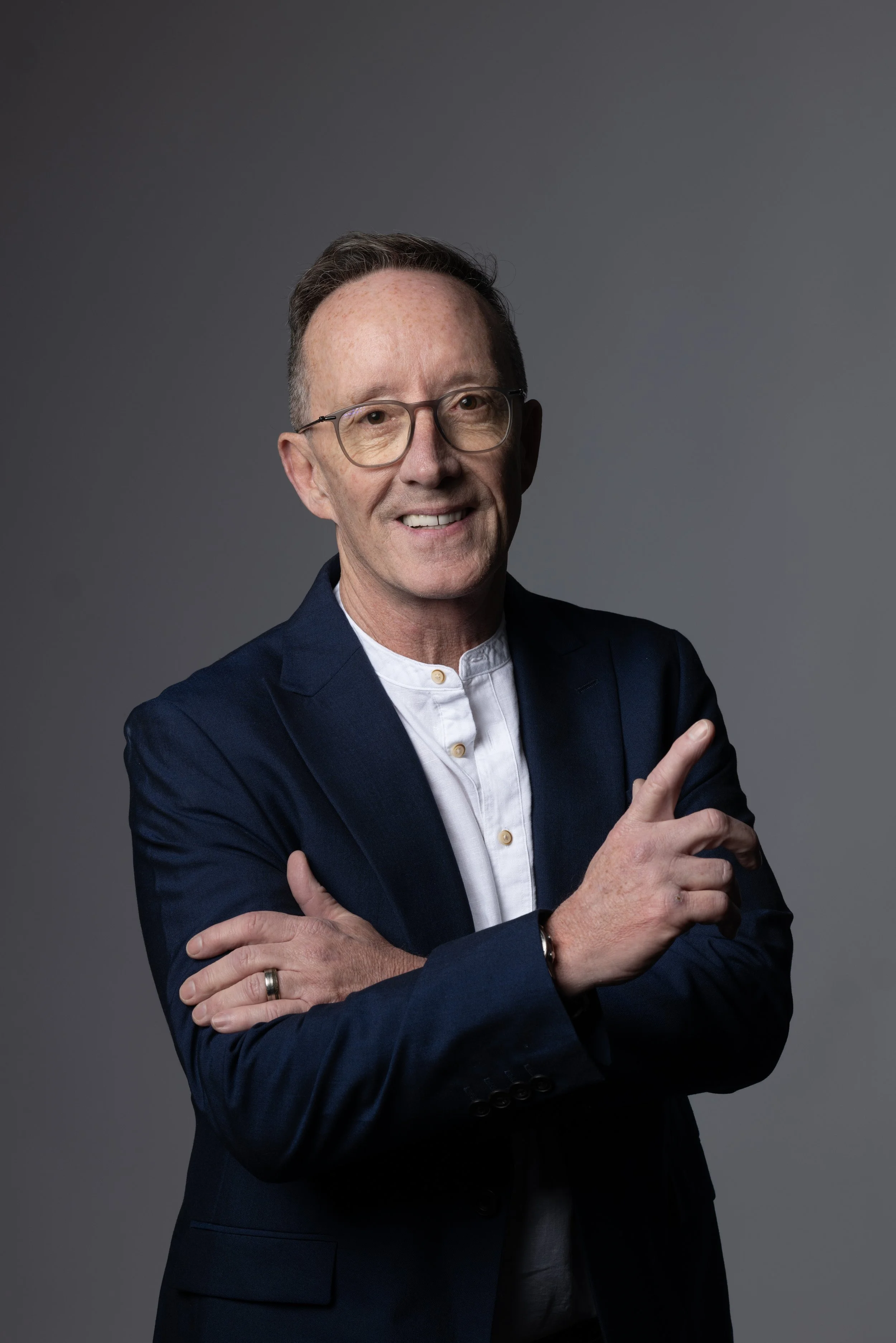 A man with glasses smiling, wearing a dark blazer and white shirt, with arms crossed and pointing upward with his right index finger.