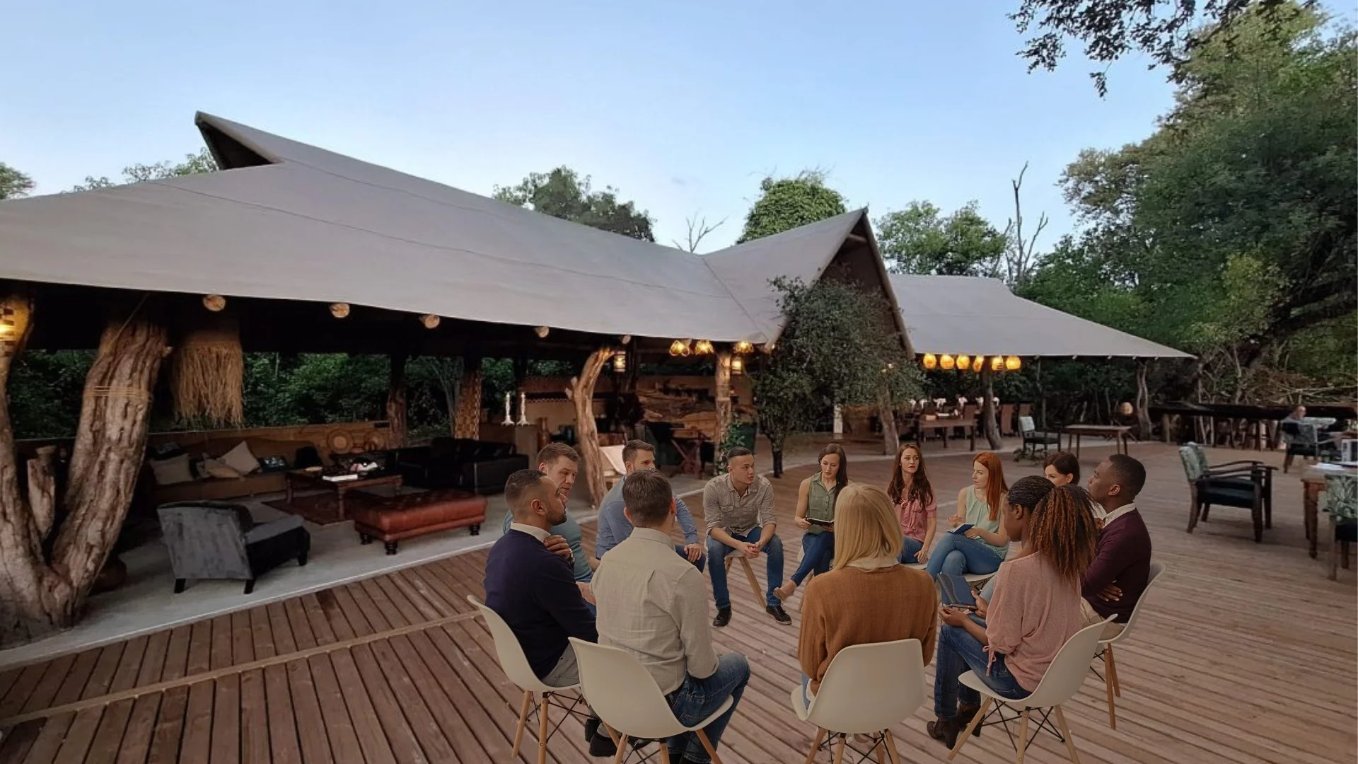 A group of people sitting in a circle outdoors on a wooden deck, engaged in conversation, with trees and a rustic building in the background.