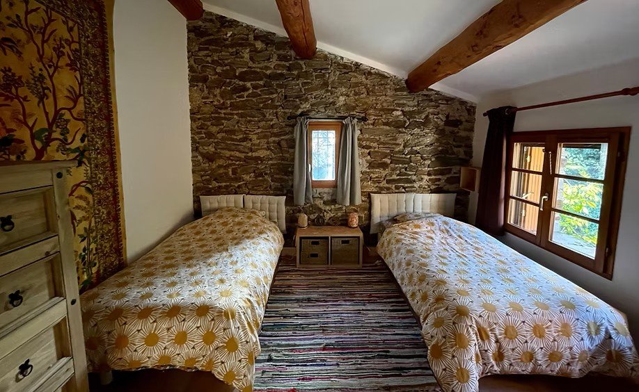 Room with two twin beds with floral bedspreads, stone wall, small window with curtains, wooden ceiling beams, large window with view outside, striped rug, and a dresser.