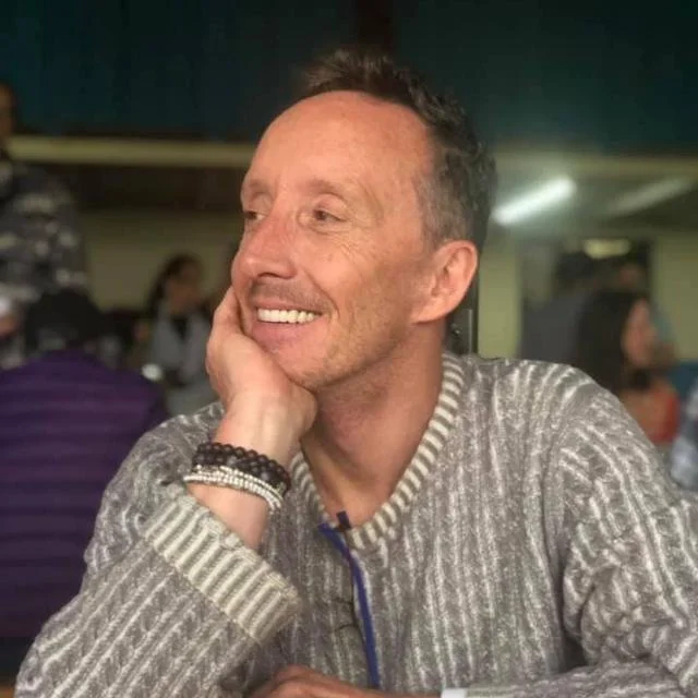 A smiling man with short dark hair and a beard, wearing a striped sweater and bracelets, resting his chin on his hand in a busy indoor setting.