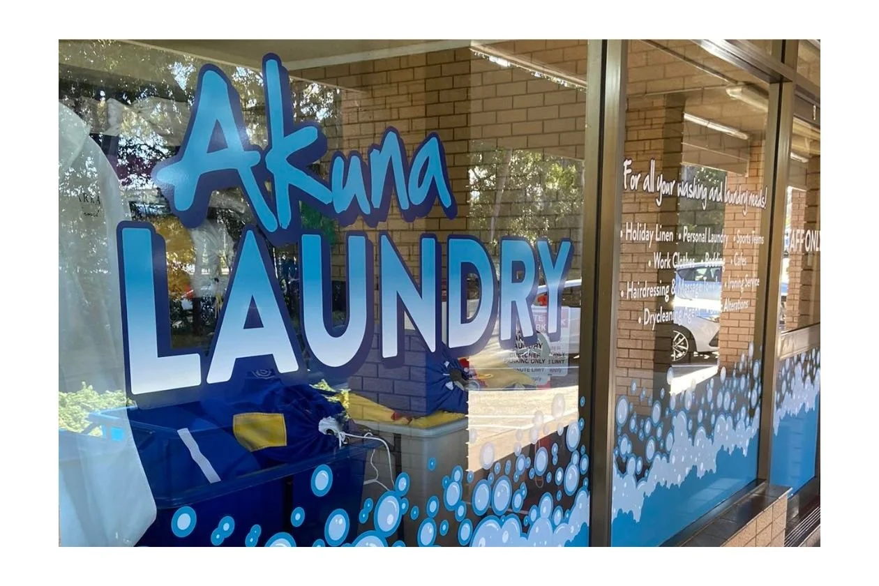 Storefront window with blue and white lettering that reads "Akuna Laundry", decorated with blue bubbles. White text lists services such as holiday linen, personal laundry, sports uniforms, work clothes, and more.