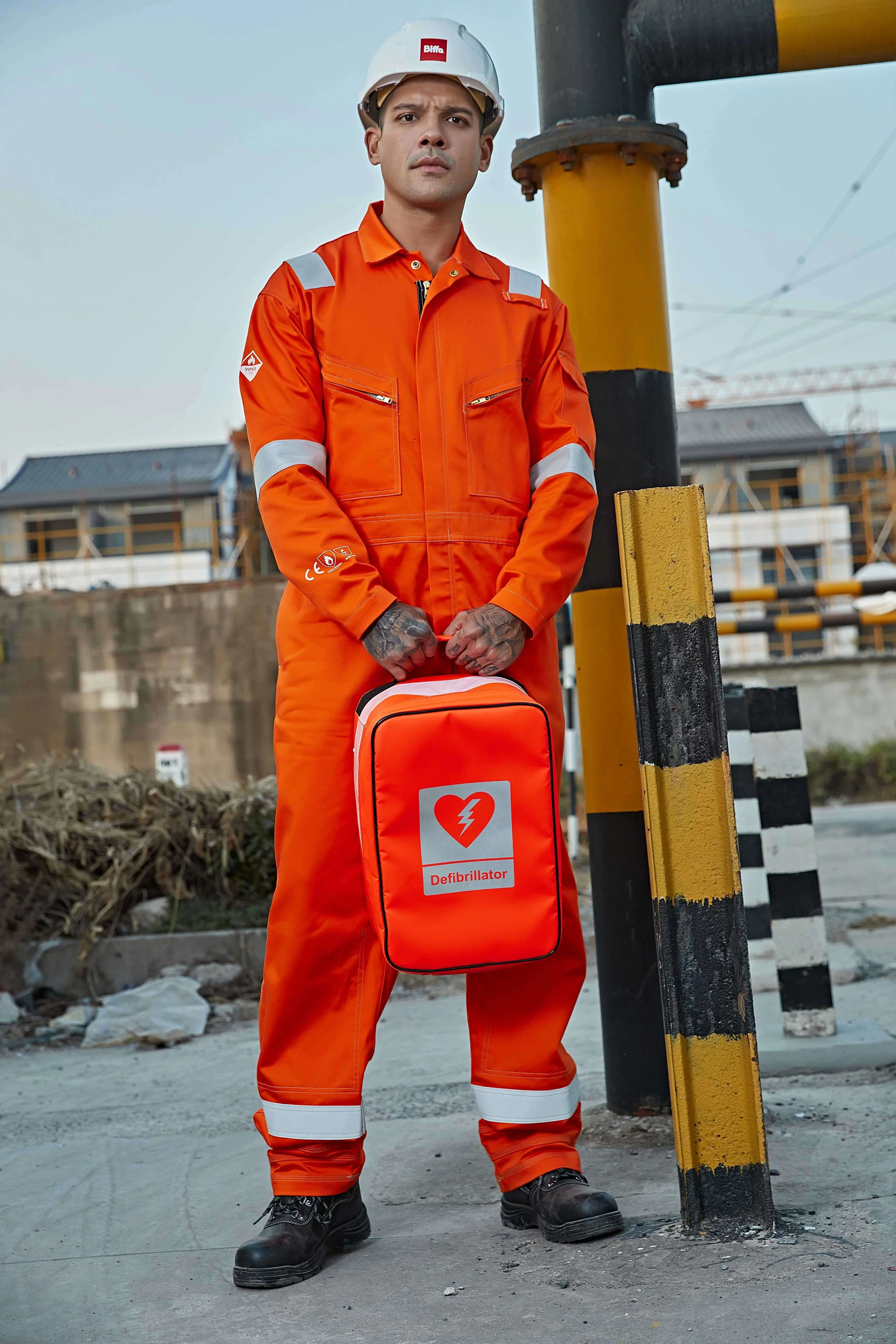 A man wearing an orange safety jumpsuit and a white hard hat, standing outdoors on a construction site, holding a LODE design bright orange defibrillator bag with a heart and lightning bolt symbol.