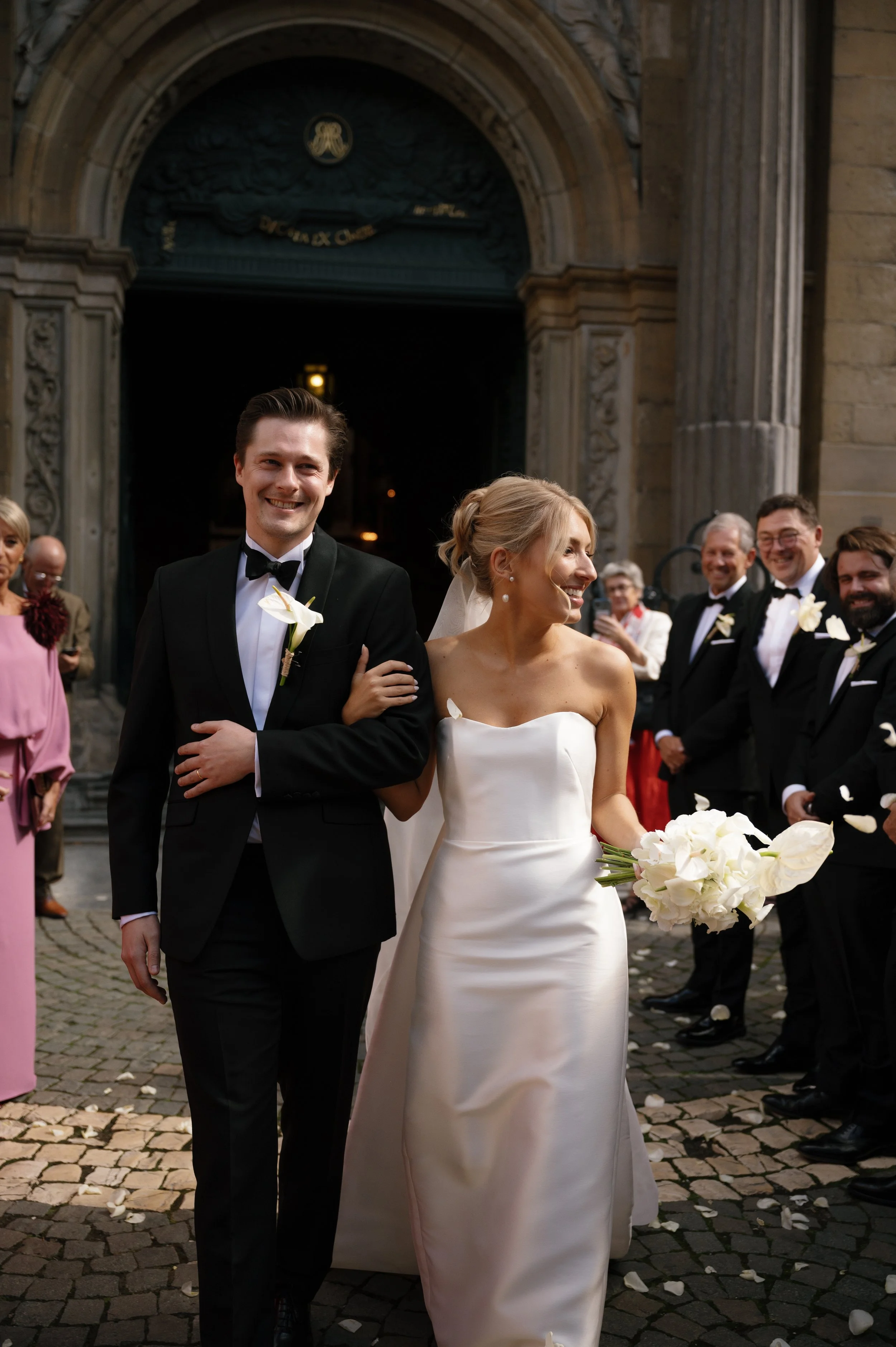 Een gelukkige getrouwde koppel loopt samen weg na de bruiloft, omringd door vrienden en familie, voor een oude kerk met een gebeeldhouwd archedoors.