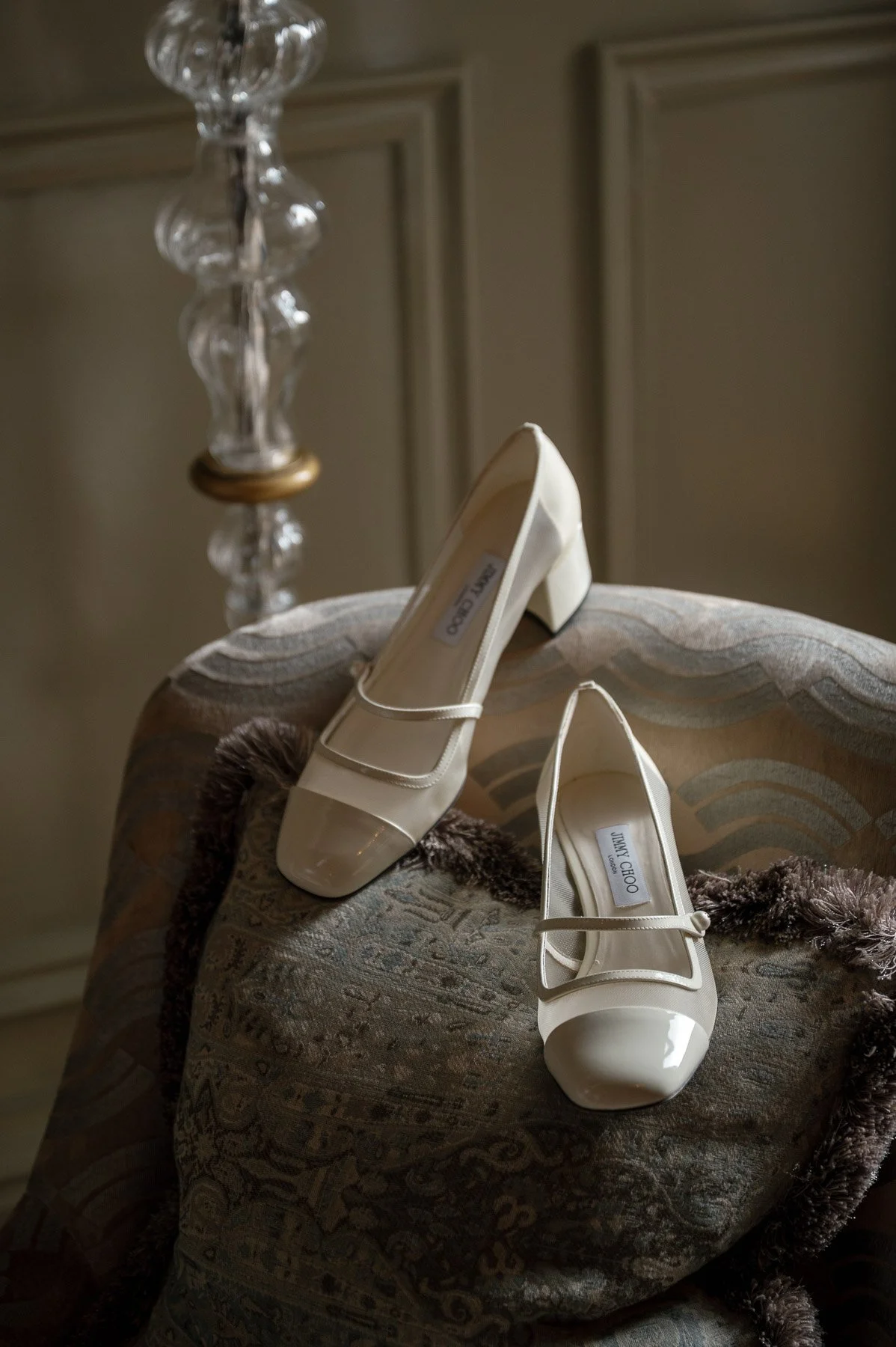 Een paar witte schoenen met lage hakken geplaatst op een gestoffeerde stoel met een donker gekleurde, pluizige rand.