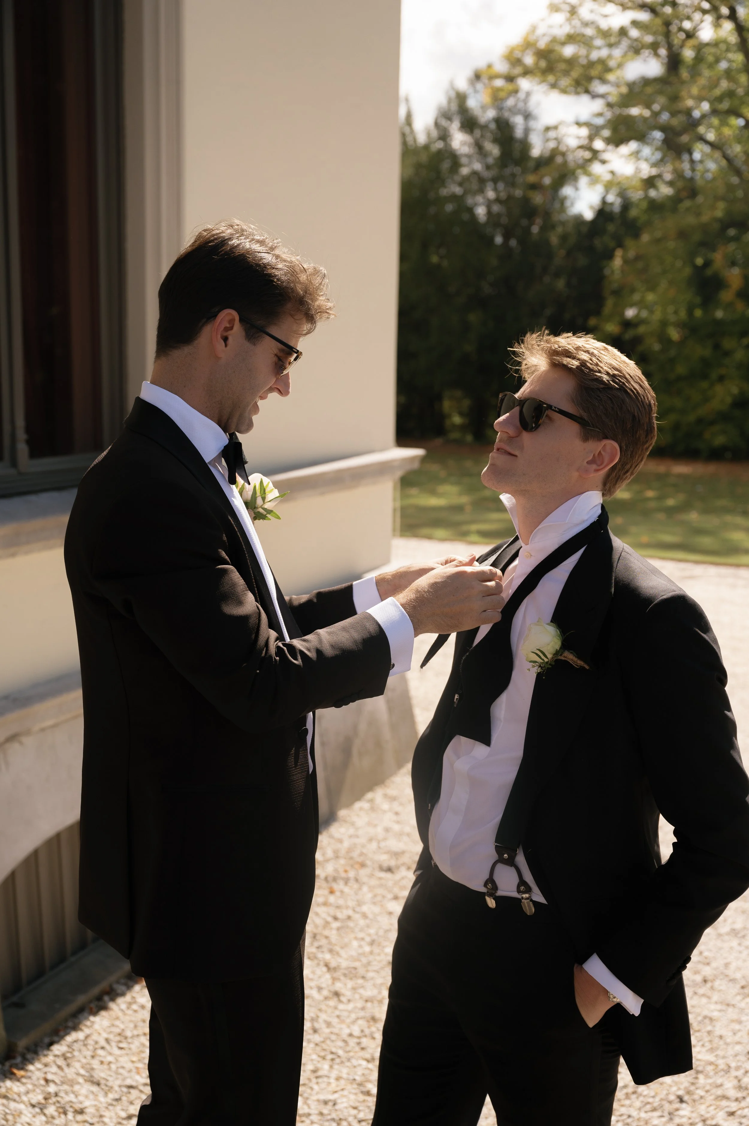 Twee mannen in smoking geven elkaar een knuffel en maken zich klaar voor een bruiloft buiten, met één man die een bloem in zijn revers heeft.