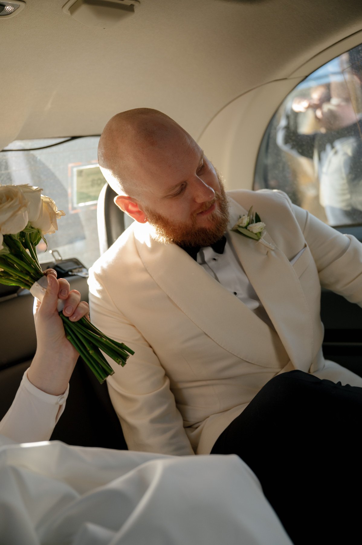 A groom with a beard in a white tuxedo jacket and black bow tie sitting on a plane seat, with a woman handing him a bouquet of white roses.