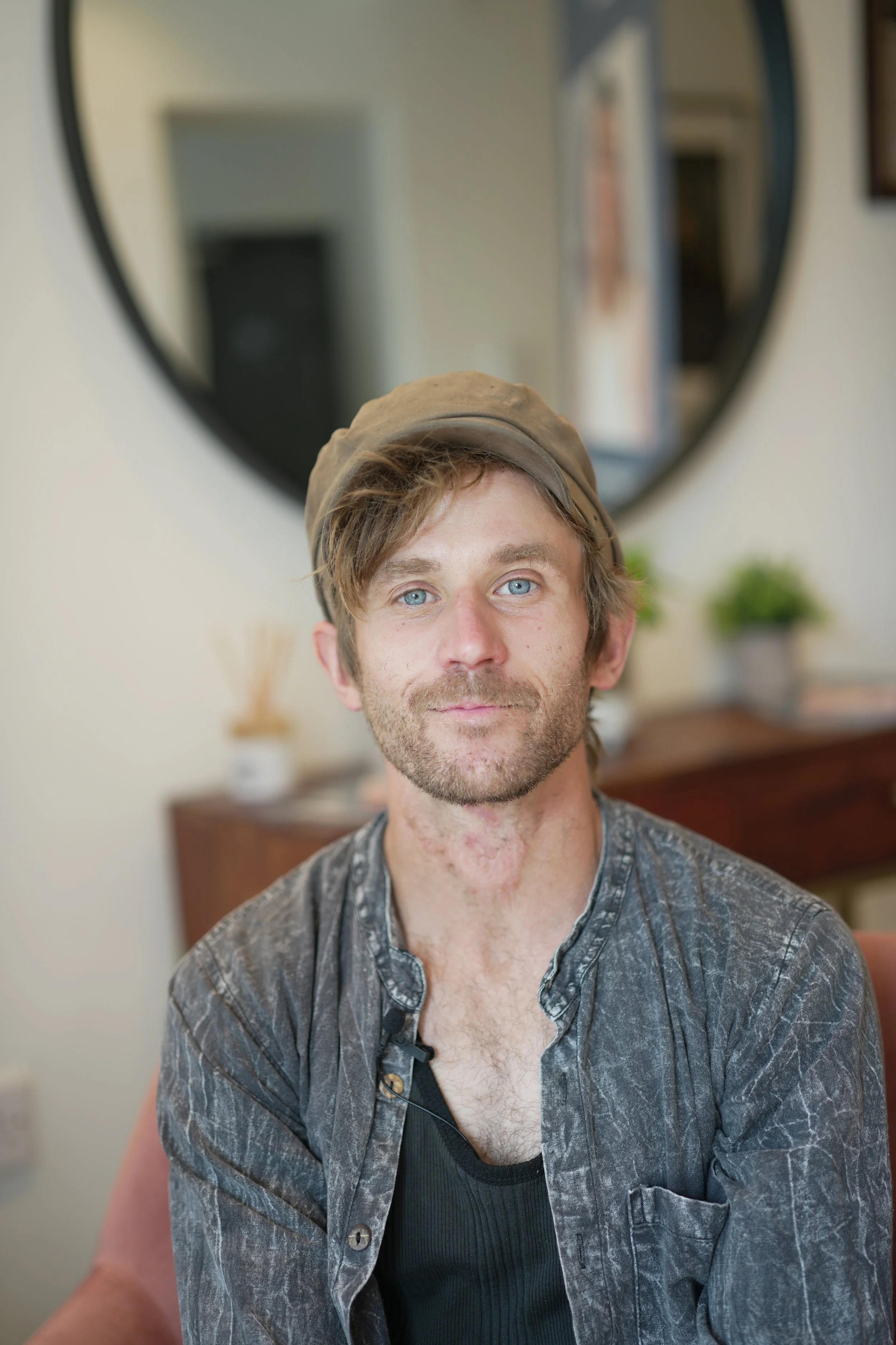A young man with light-colored hair and blue eyes wearing a beige cap, black tank top, and a grey open button-up shirt, sitting at a desk in a room with a large round mirror and blurred background.