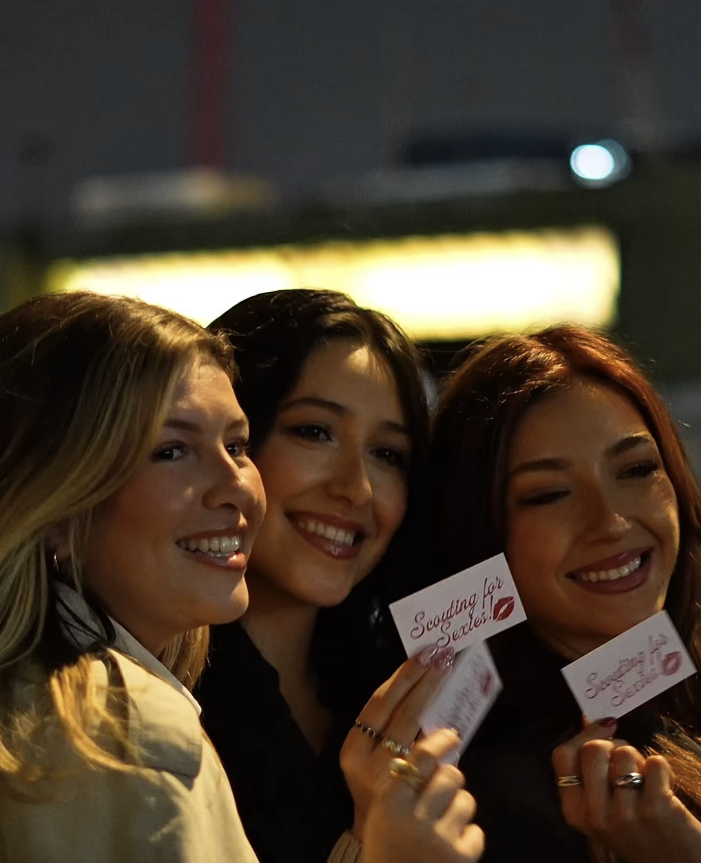 Three women smiling and holding 'Scouting for Secrets' cards in a dimly lit setting.