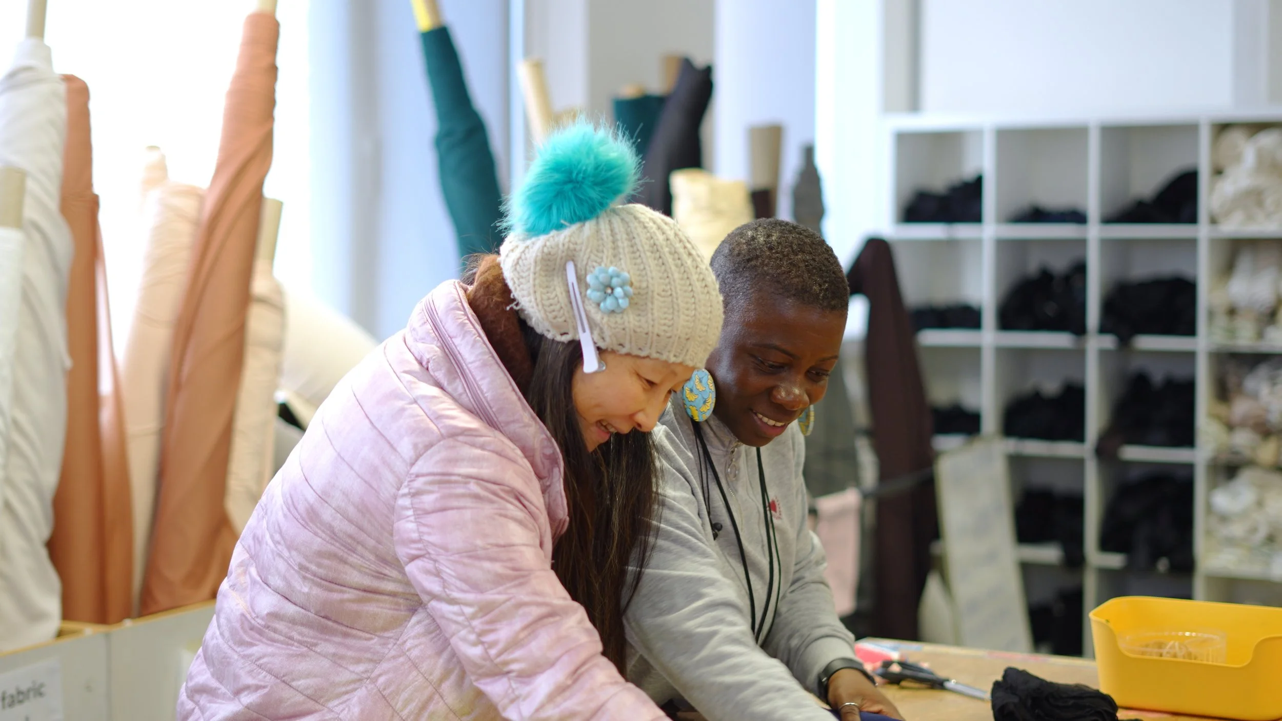 Two women working together at a crafting or sewing project in a craft store or workshop, with shelves containing fabric in the background.