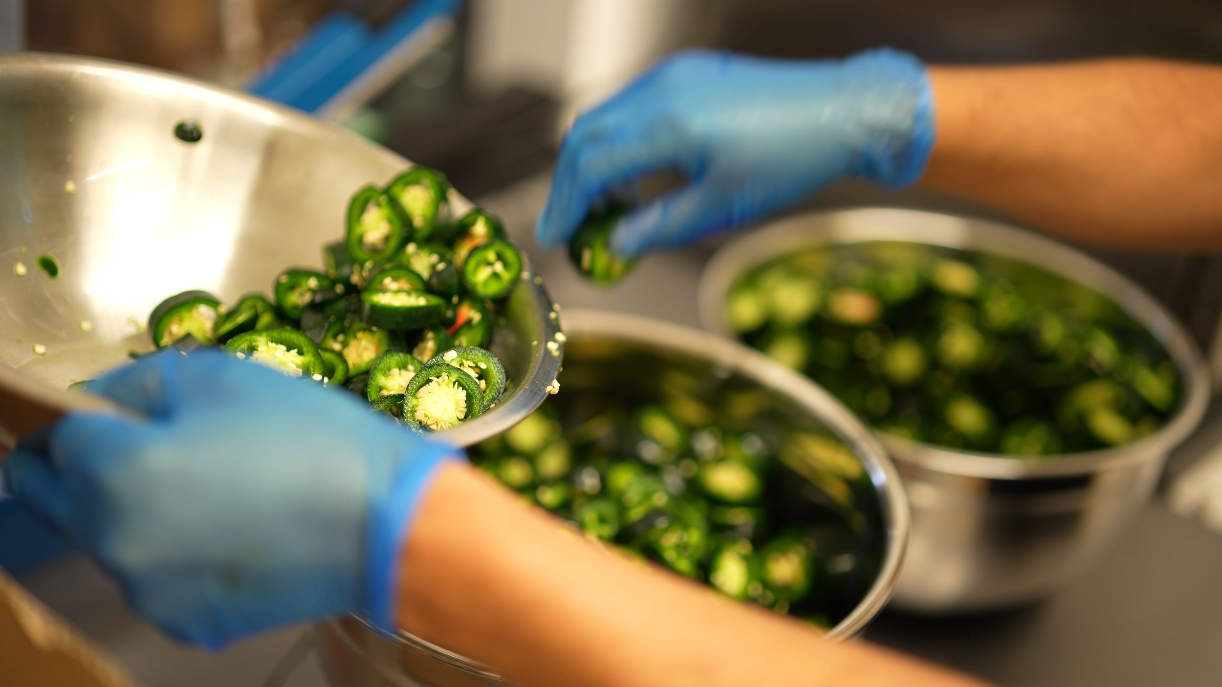 A person wearing blue gloves slicing fresh green jalapeño peppers over a metal bowl, with more peppers in the background.