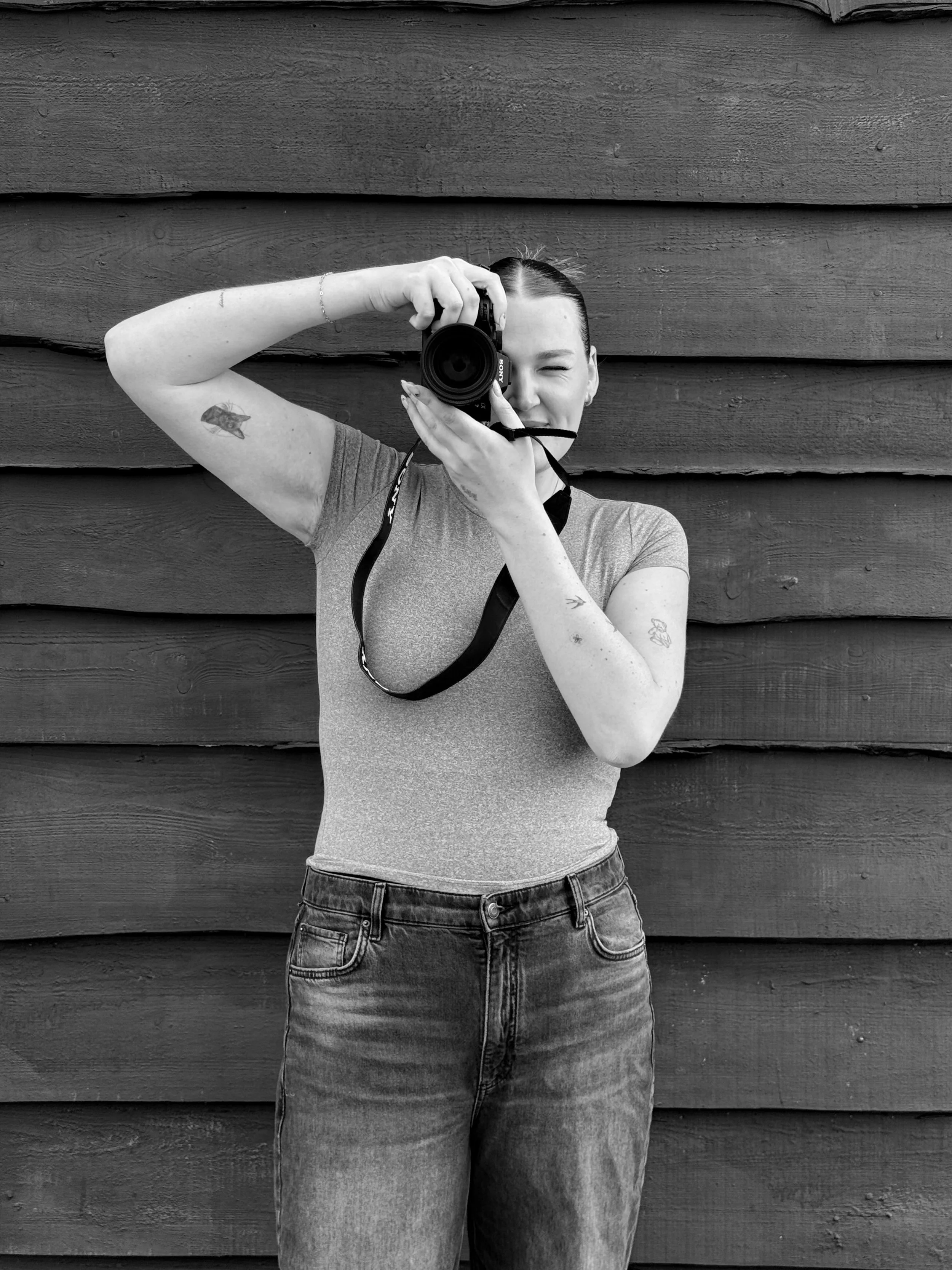 A young woman with dark hair in a ponytail, wearing a short-sleeved shirt and jeans, taking a mirror selfie with a camera, standing against a wooden wall, smiling and winking, with tattoos on her left arm.