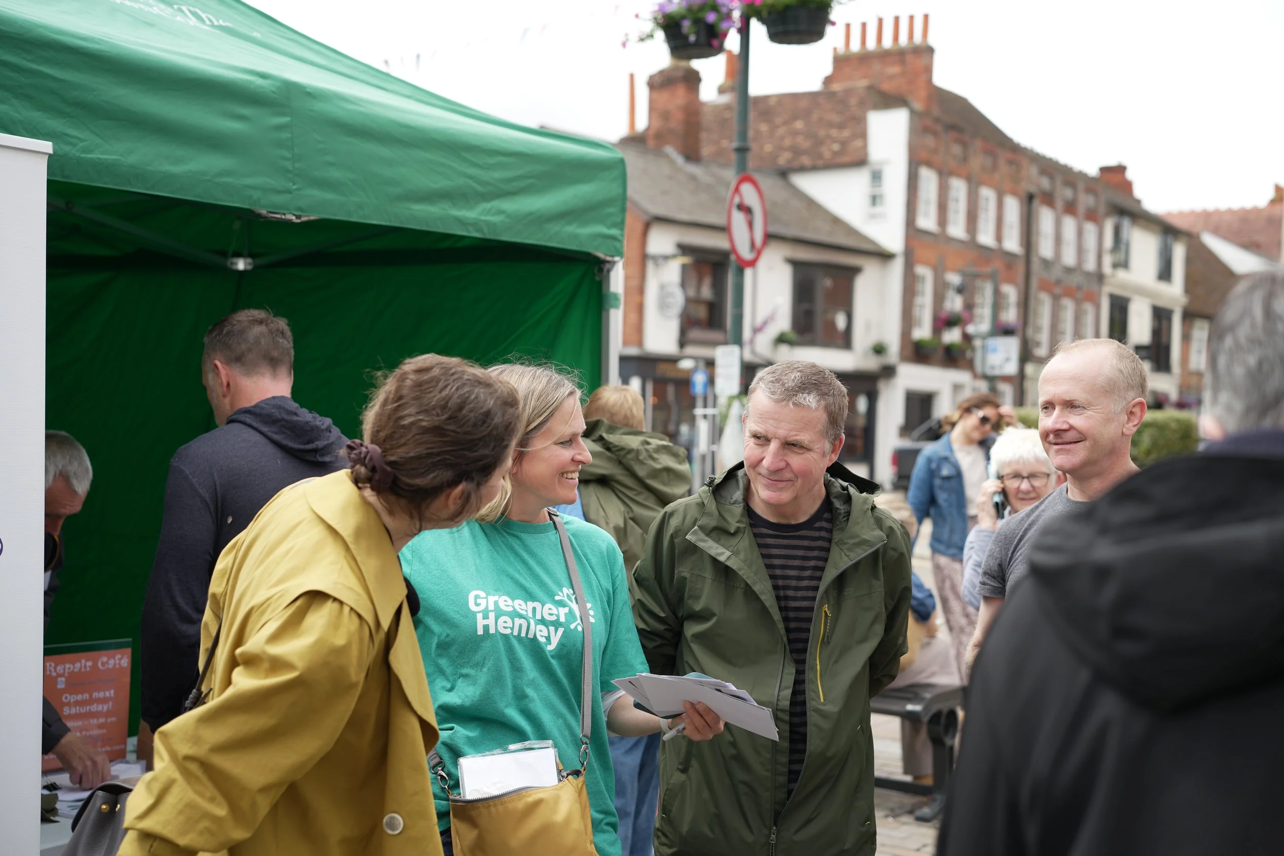 Group of people at an outdoor event, with a woman in a green shirt and yellow jacket talking to a man in a green jacket. Other individuals are gathered around, engaging in conversation, with booths and buildings visible in the background.
