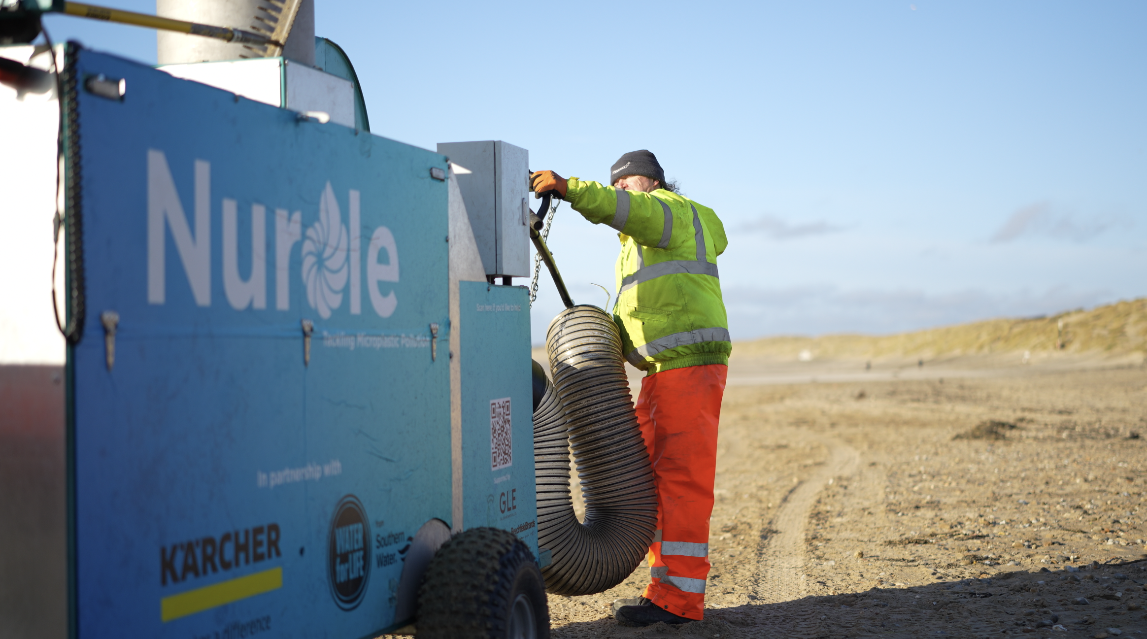 A worker in a bright yellow safety jacket, orange pants, and a gray beanie working on a large blue filtration unit labeled "Nurelle" in an outdoor area with dirt ground and a clear sky.