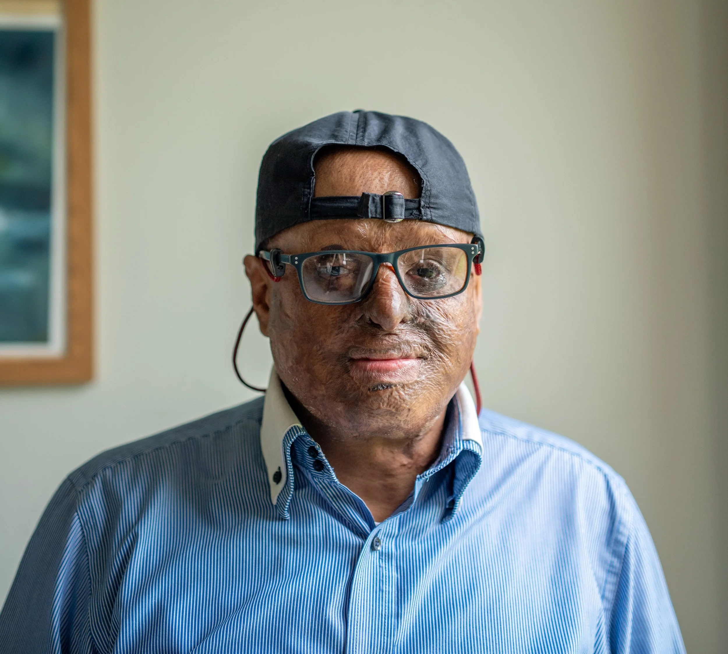 Close-up of an elderly man wearing glasses, a black cap worn backward, and a blue striped shirt.