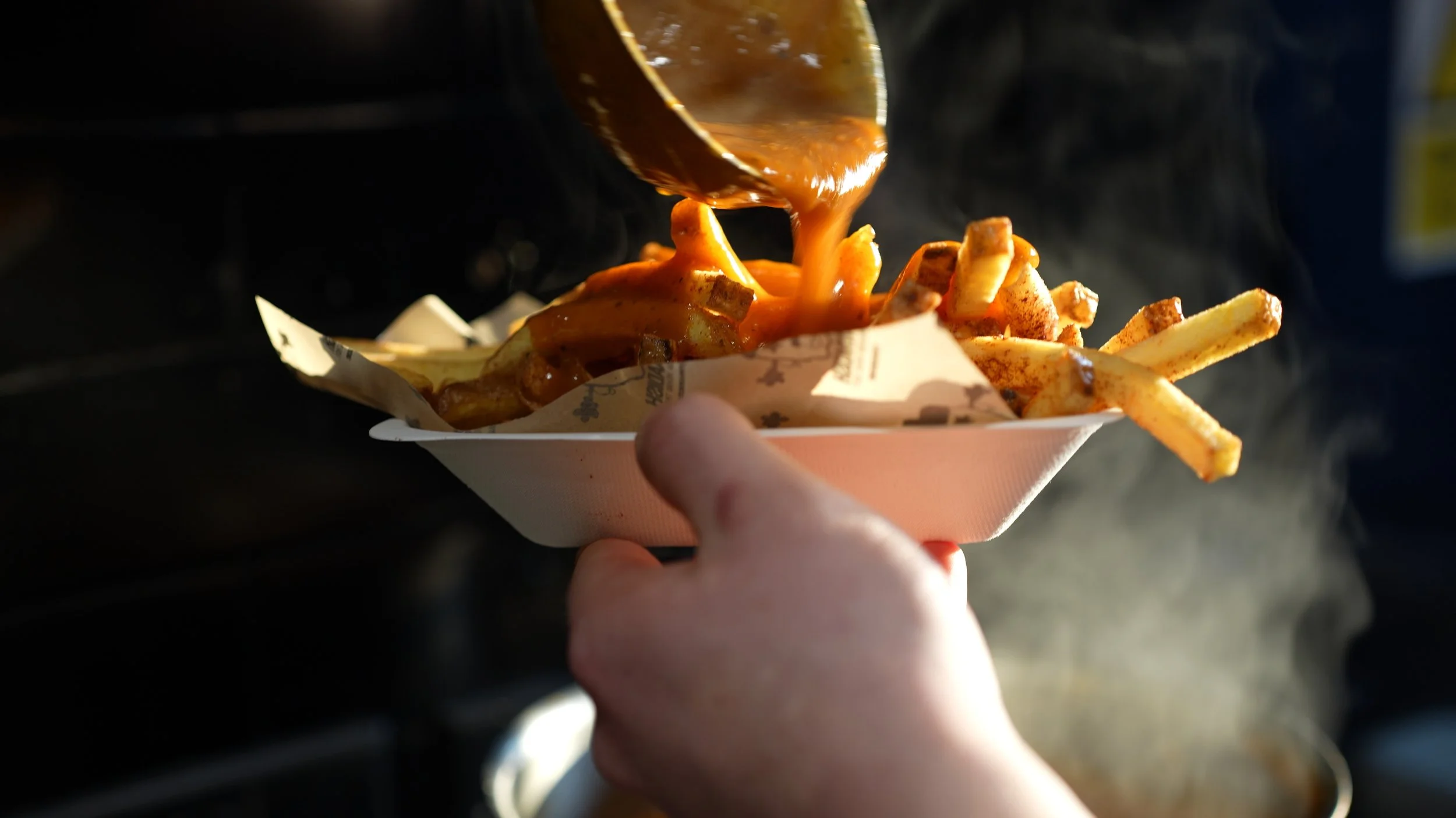 Fried French fries topped with chili and cheese, served in a paper food tray, with a person holding it.