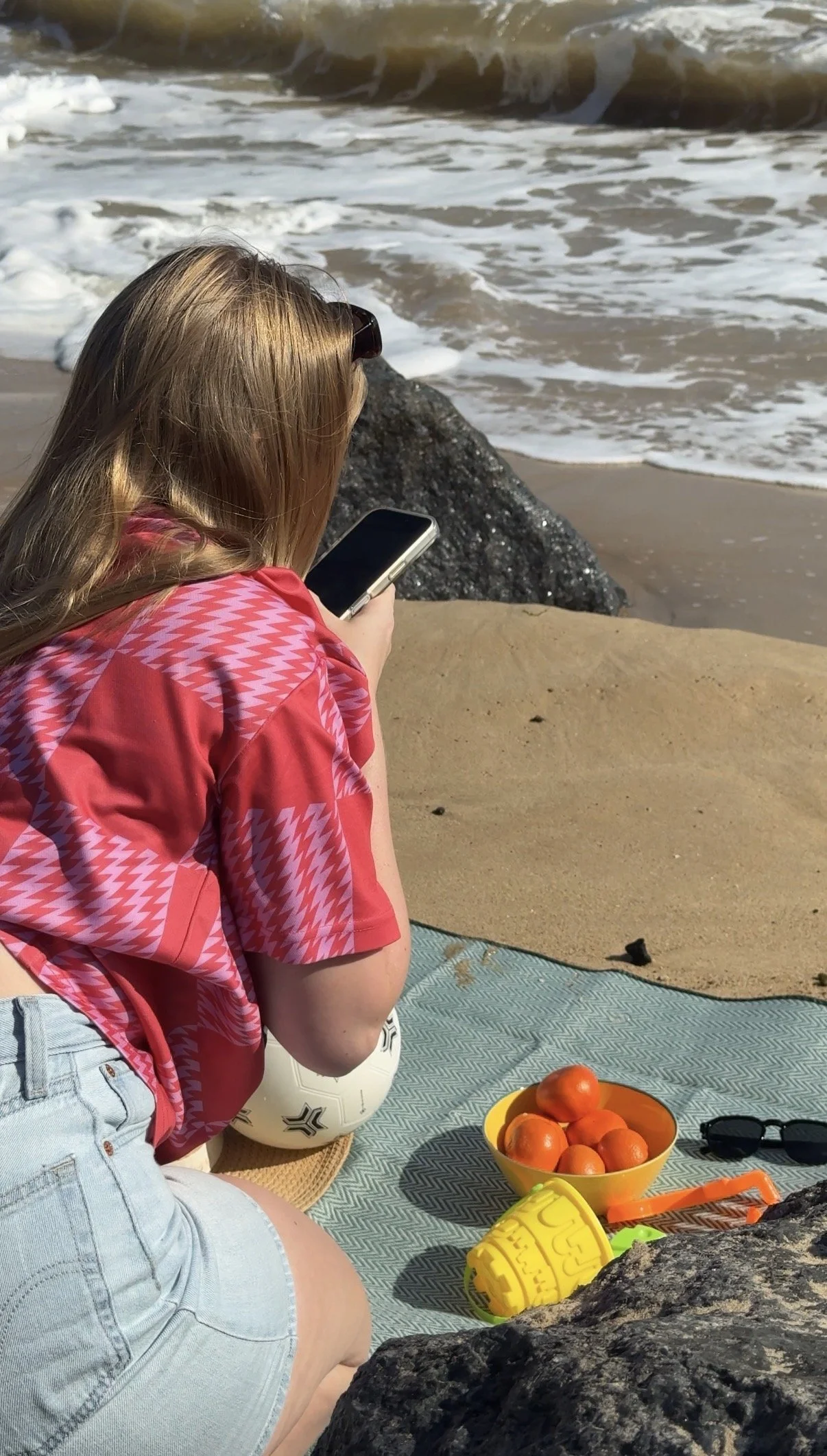 A woman with light brown hair wearing a red and pink checkered shirt and denim shorts sitting on a beach blanket, looking at her phone near the sandy shore with waves in the background. A yellow bowl of oranges and some toys are on the blanket.