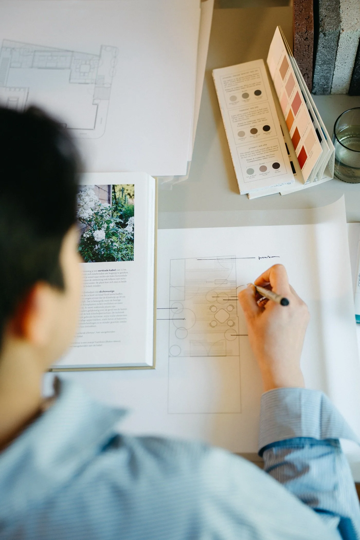 Een vrouw van Studio Jong Baw schetst een tuinontwerp voor een ecologische stadstuin, en gebruikt daarbij kleurstalen van Farrow & Ball, een boek over planten en materialen ter inspiratie