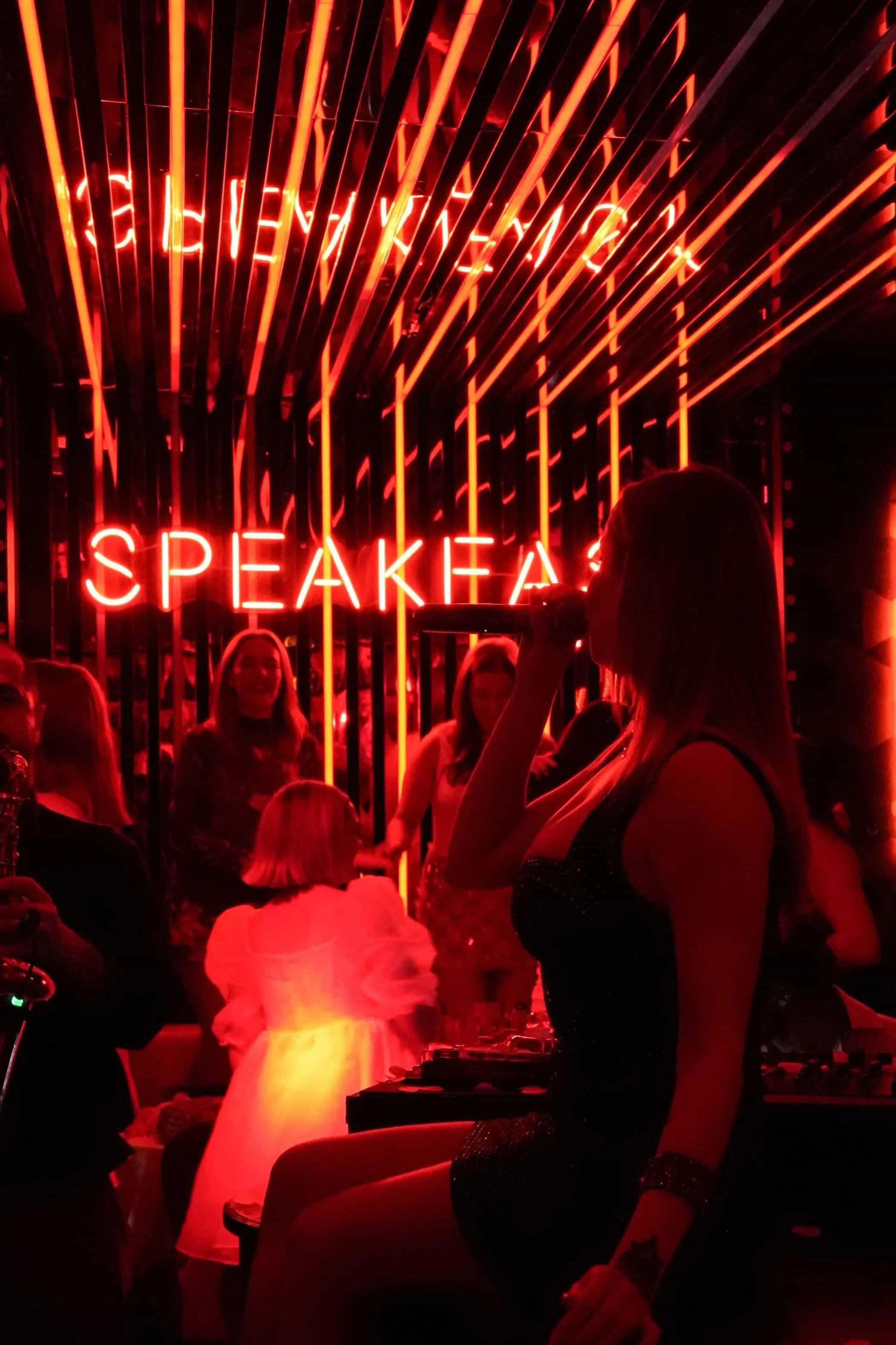 Une femme chante dans un club avec un microphone, entourée d'autres personnes, avec un panneau lumineux rouge affichant le mot 'SPEAK' en arrière-plan.