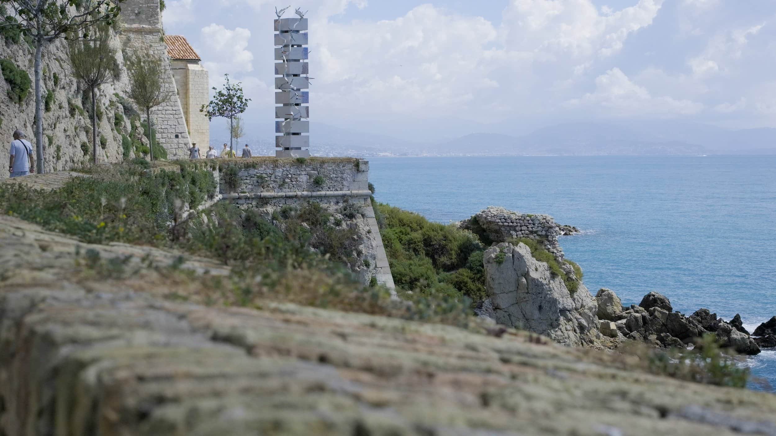 Vue de la côte méditerranéenne avec une promenade en pierre, des rochers, une mer bleue, un ciel partiellement nuageux et une sculpture moderne en arrière-plan.