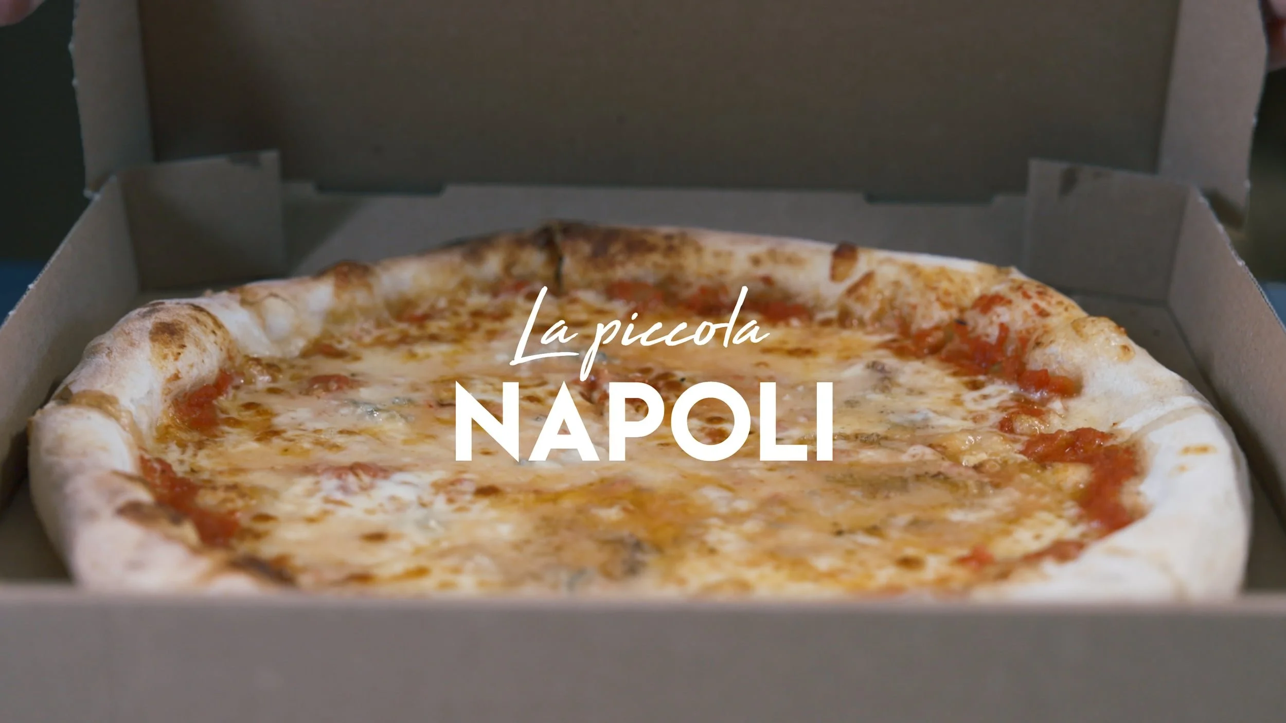 Une pizza dans une boîte à pizza ouverte, garnie de fromage, tomate et autres ingrédients, avec le texte "La piccola NAPOLI" affiché dessus.