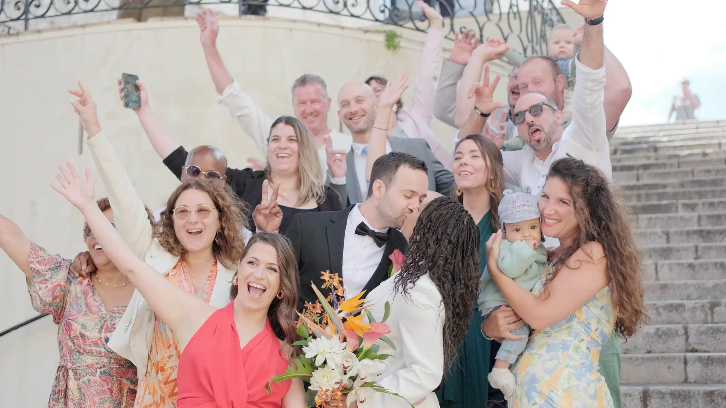 Groupe de personnes joyeuses posant pour une photo de mariage, avec un couple d'amoureux s'embrassant au centre, entourés d'amis et de famille sur des escaliers.