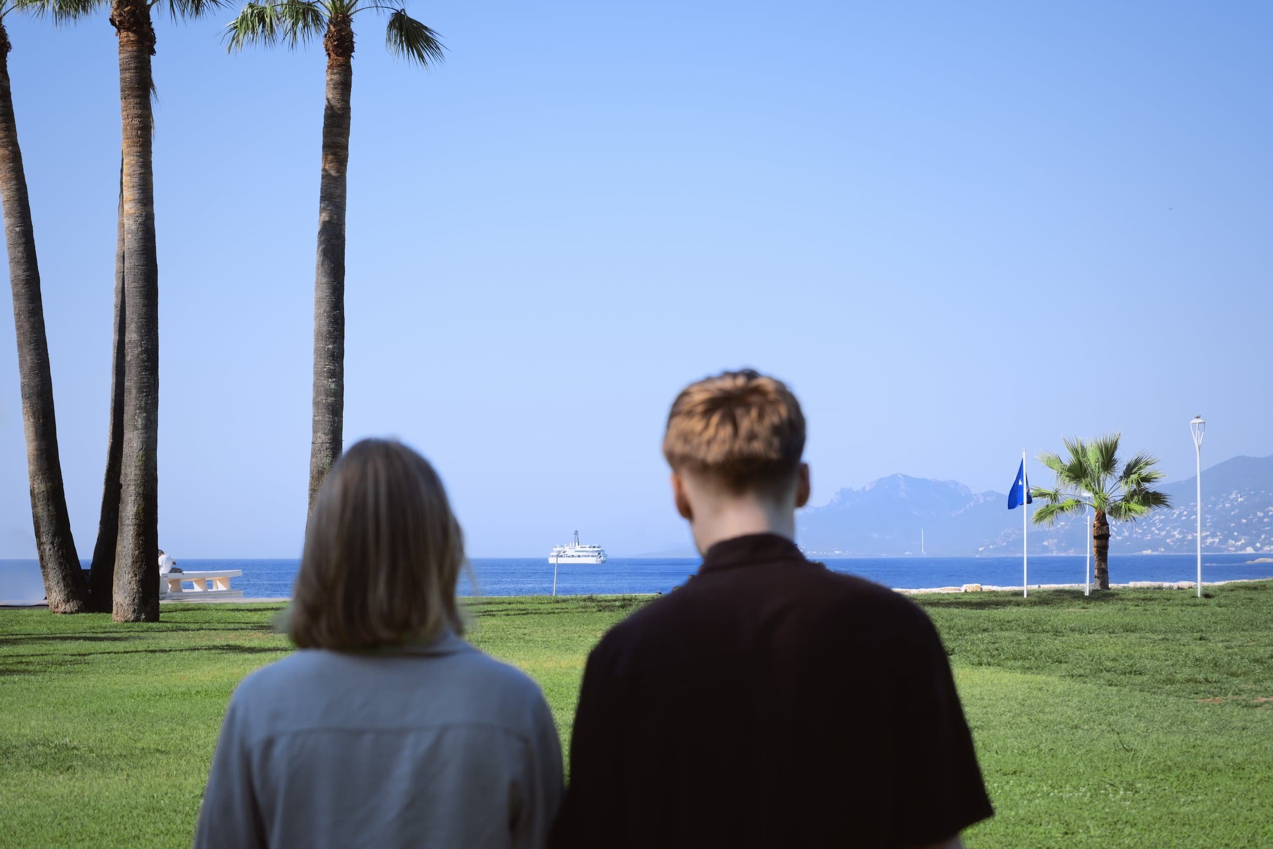 Deux personnes regardant la mer depuis un parc avec des palmiers et un bateau au loin.