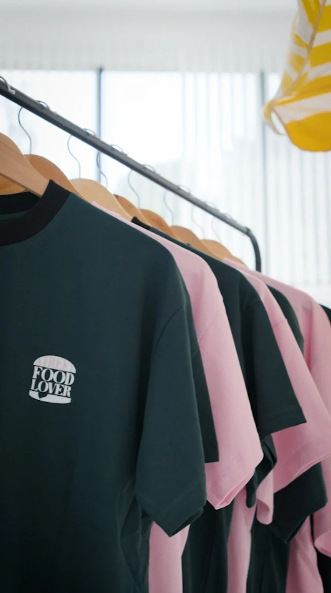 Groupe de t-shirts noirs et roses suspendus sur une tringle dans un magasin, avec un logo "FOOD LOVER" sur un t-shirt noir en premier plan.
