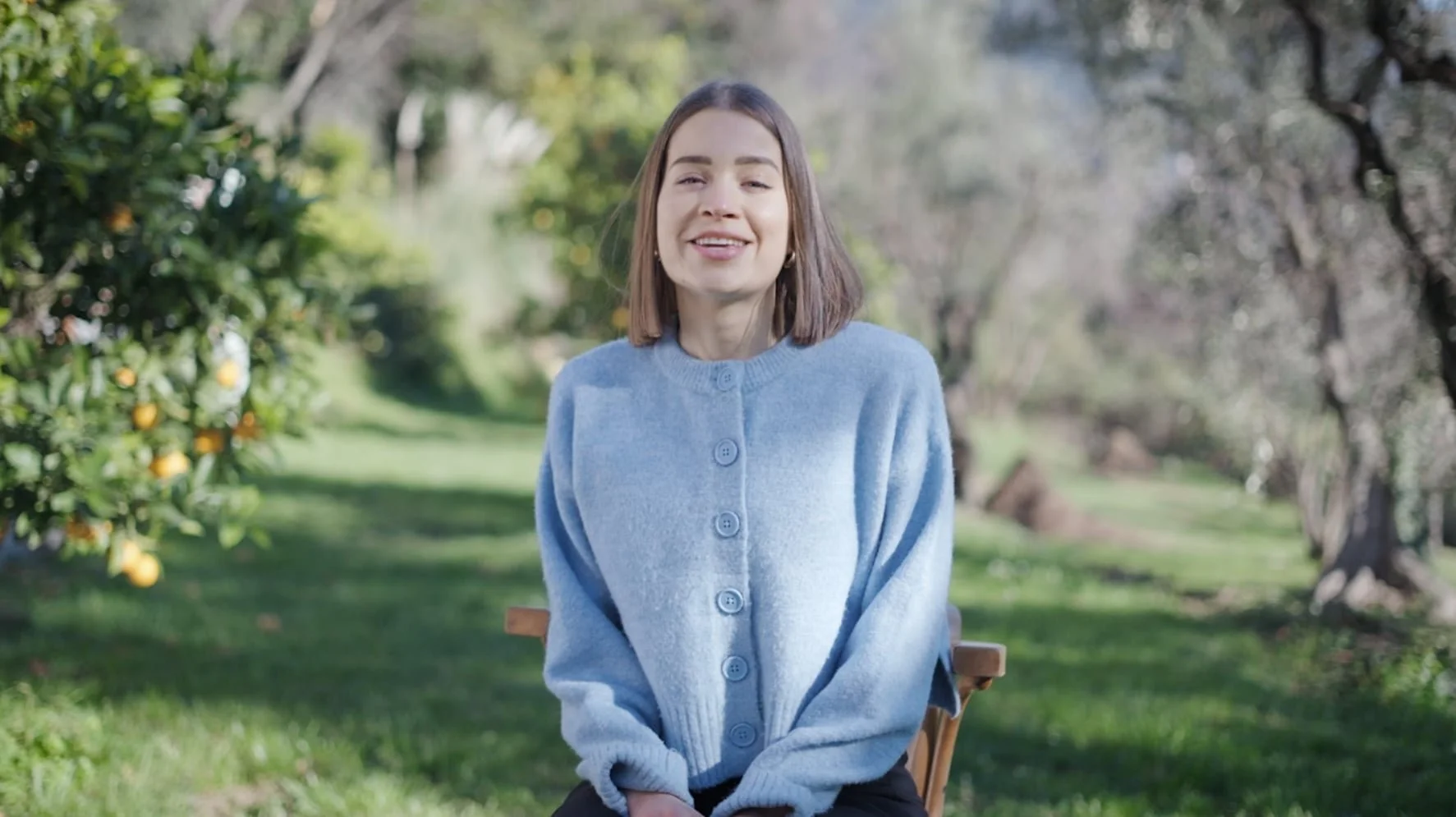 Femme souriante assise sur une chaise dans un jardin avec des arbres et des citrus, vêtue d'un pull bleu clair.