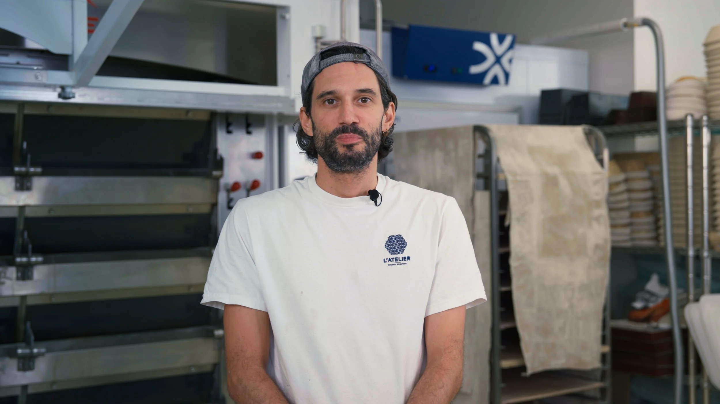 Un homme avec une barbe, portant un t-shirt blanc avec un logo L'ATELIER, se tenant dans un atelier, avec des étagères en métal et divers matériaux autour.