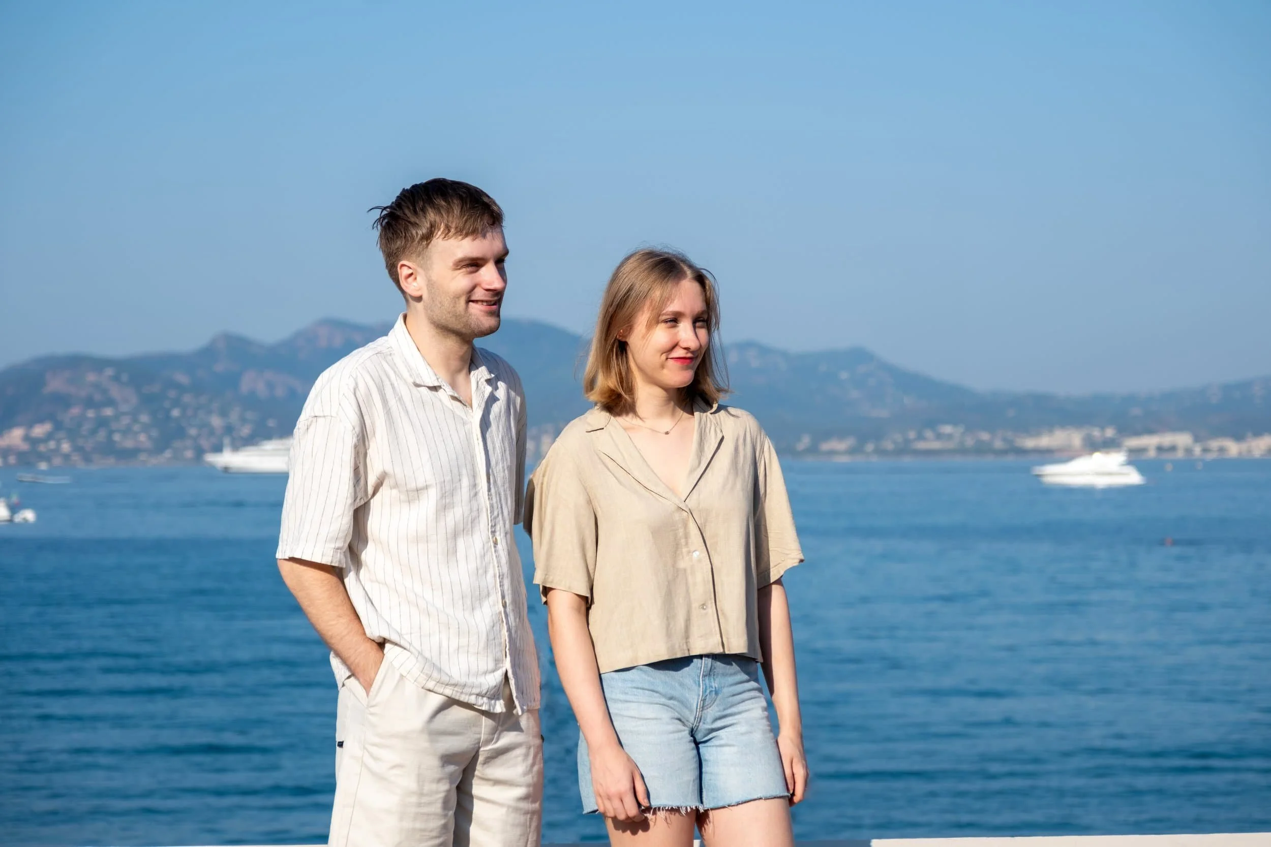 Un jeune homme et une jeune femme debout près de l'eau avec des montagnes en arrière-plan, rions et regardent vers la droite.
