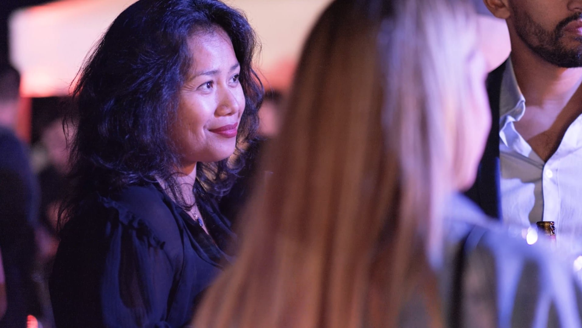 Une femme souriante avec des cheveux bouclés, lors d'un événement social ou professionnel, entourée d'autres personnes. L'ambiance est chaleureuse avec un éclairage tamisé.
