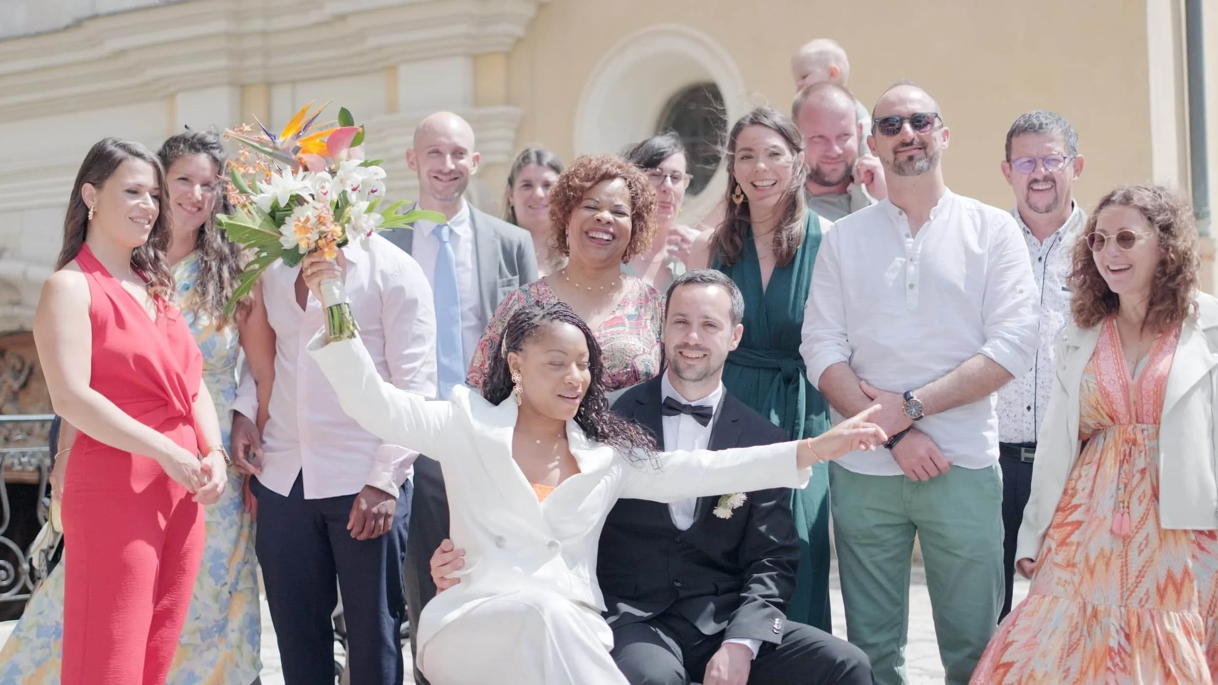 Groupe de personnes célébrant un mariage en plein air, avec la mariée et le marié au centre, la mariée tenant un bouquet de fleurs.