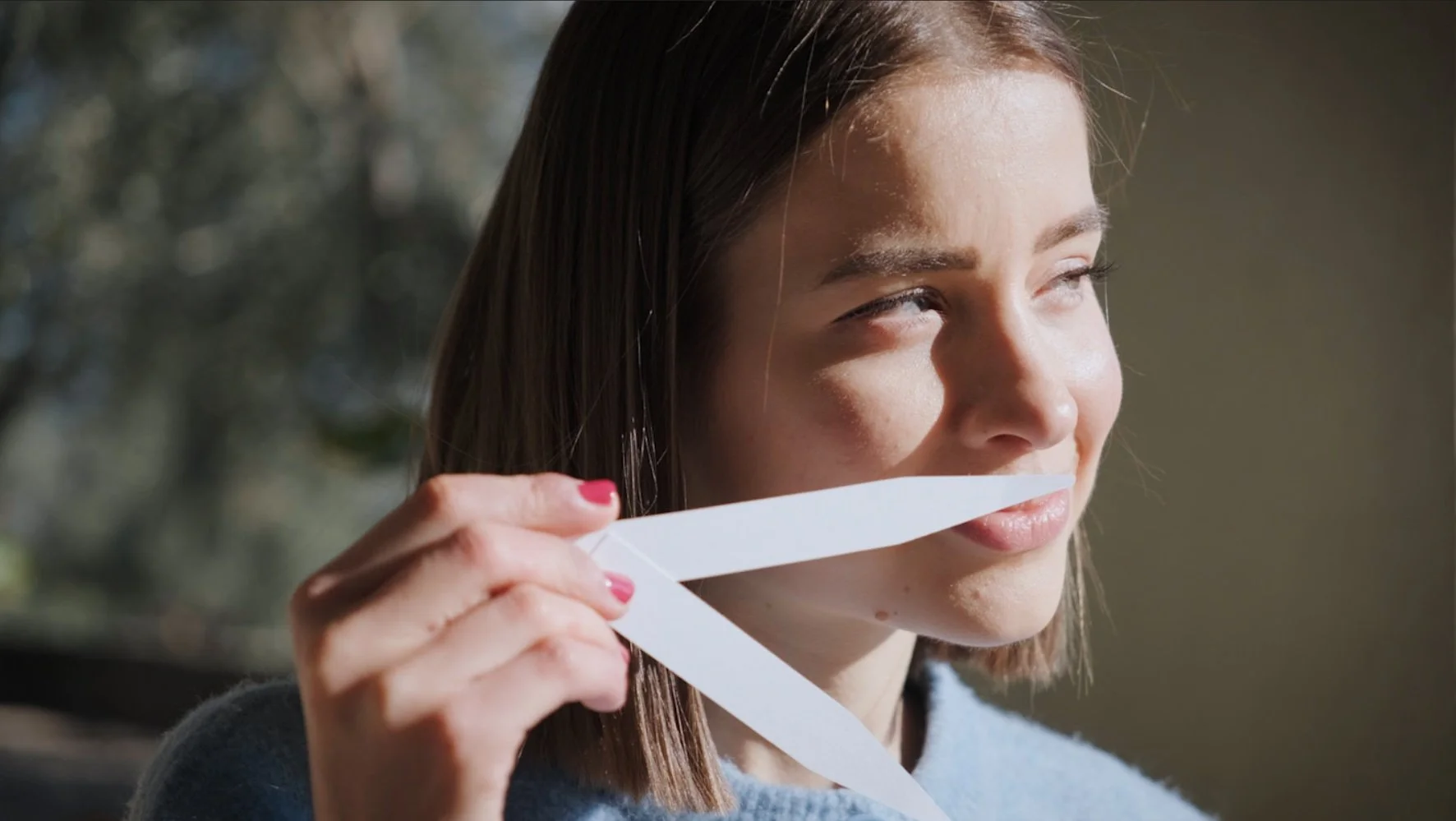 Jeune femme avec des cheveux bruns clair tenant une bande de papier blanc près de sa bouche, regardant à droite, avec un arrière-plan flou et un éclairage naturel.