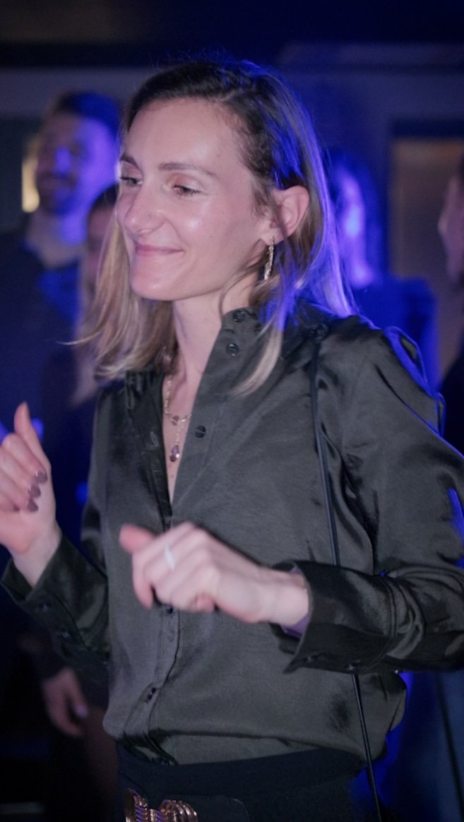 Femme souriante et dansant dans un endroit sombre avec un éclairage bleu, portant une veste en cuir noire et des bijoux.