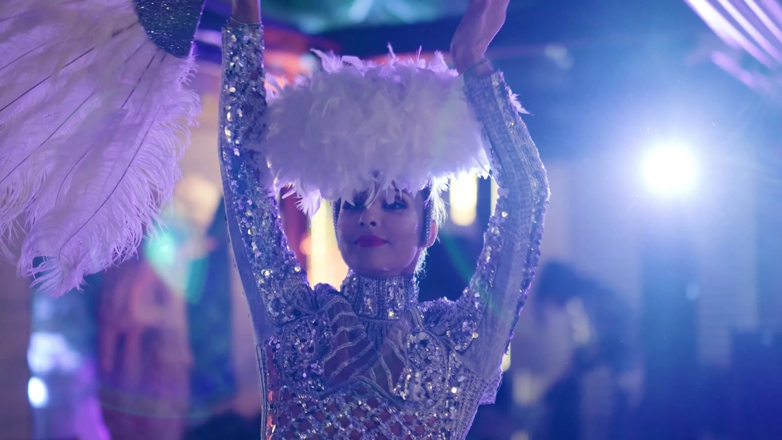 Une femme portait une robe scintillante avec des paillettes et un collier, portant un masque ou un accessoire en plumes blanches au-dessus de la tête lors d'une performance ou d'une fête, avec un éclairage coloré en arrière-plan.