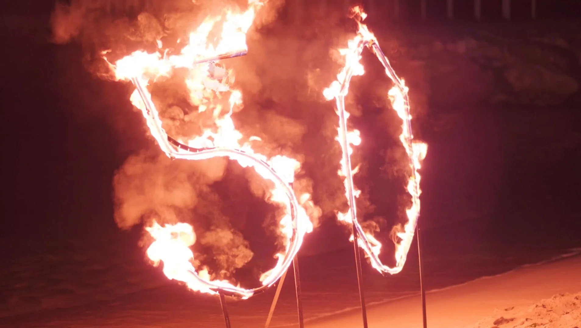 Une structure en forme de lettres enflammées située sur une plage, avec la mer en arrière-plan la nuit.