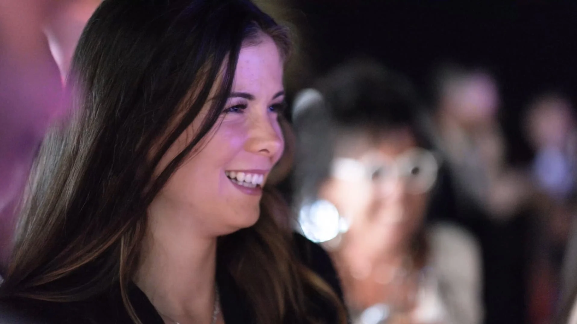Une femme souriante, avec des cheveux bruns, lors d'une fête ou d'un événement social, entourée d'autres personnes floues en arrière-plan.